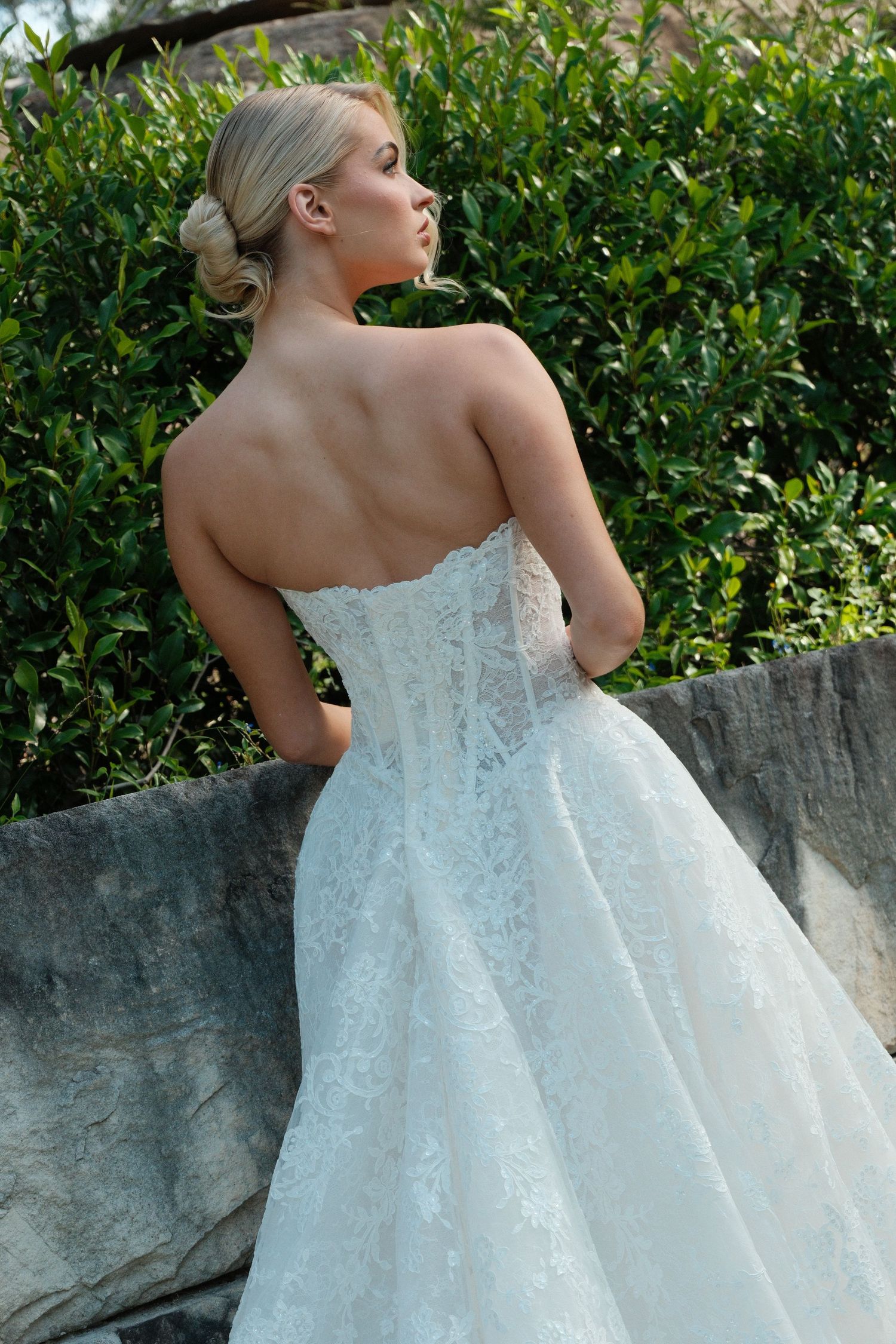 A bride in a white lace wedding gown with a flowing skirt poses outdoors against a garden wall.