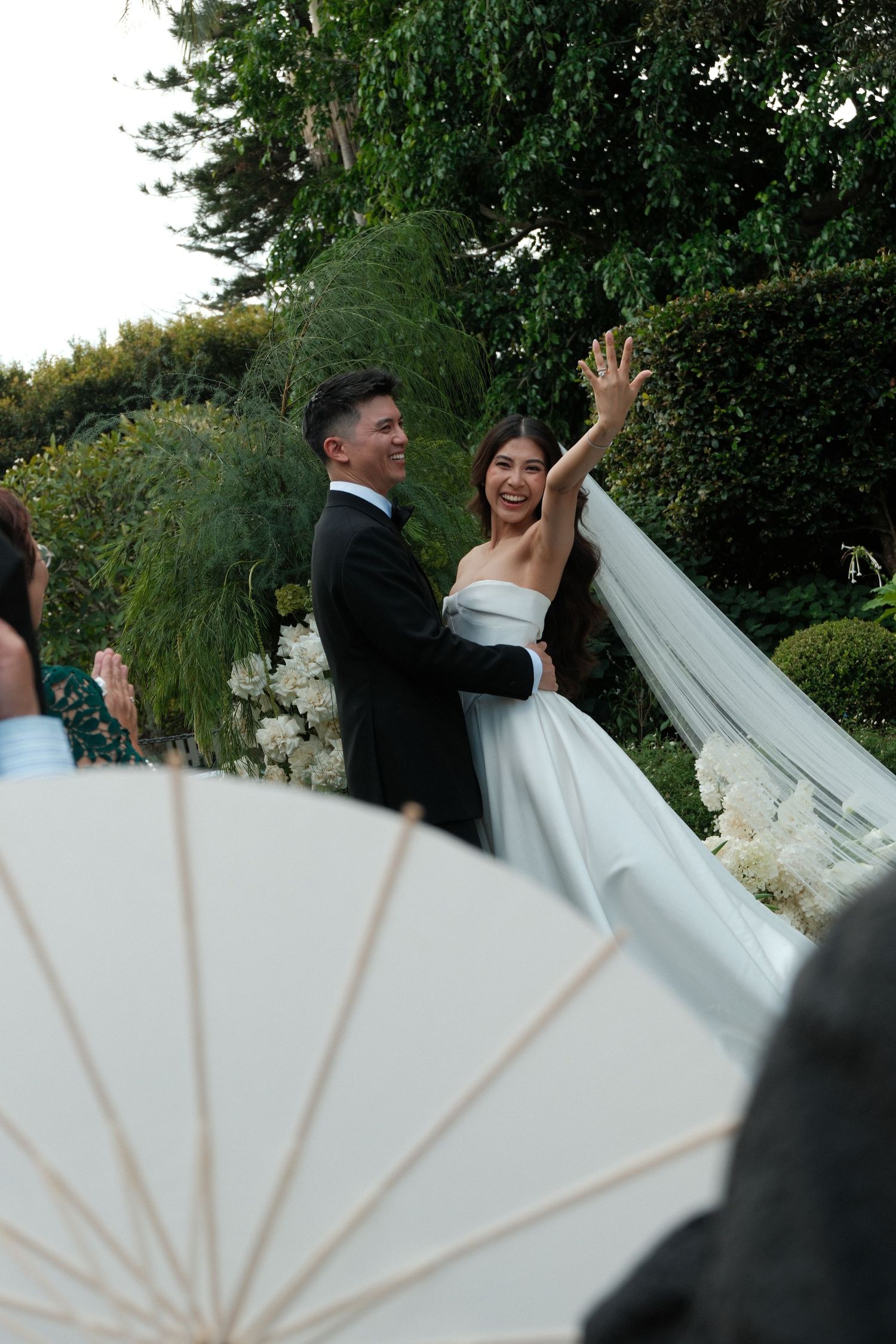 A bride and groom dance together outdoors with a decorative white paper fan in the foreground.