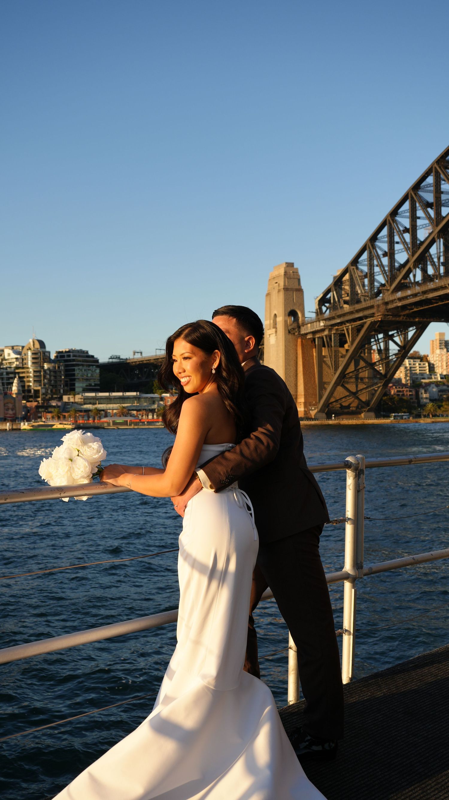 Bride and groom embrace on Sydney Harbour waterfront with iconic Harbour Bridge backdrop on sunny wedding day.