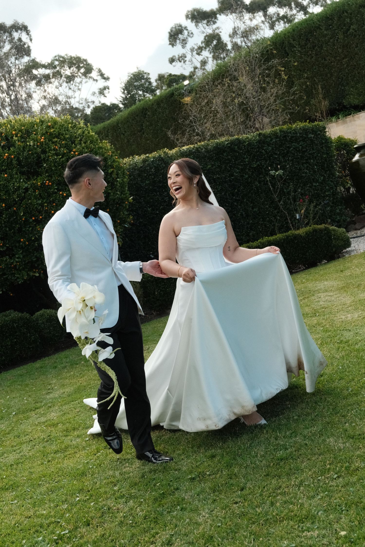 Happy bride in white strapless gown lifts dress while laughing with groom in white tuxedo jacket on green lawn.