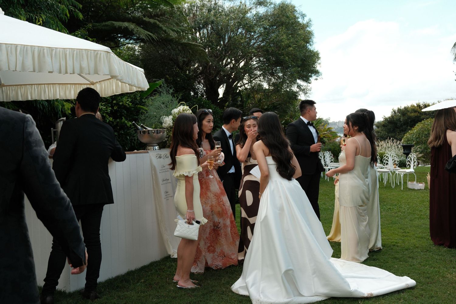 Wedding guests gather at an outdoor reception bar area with white umbrellas and green lawn in the background.