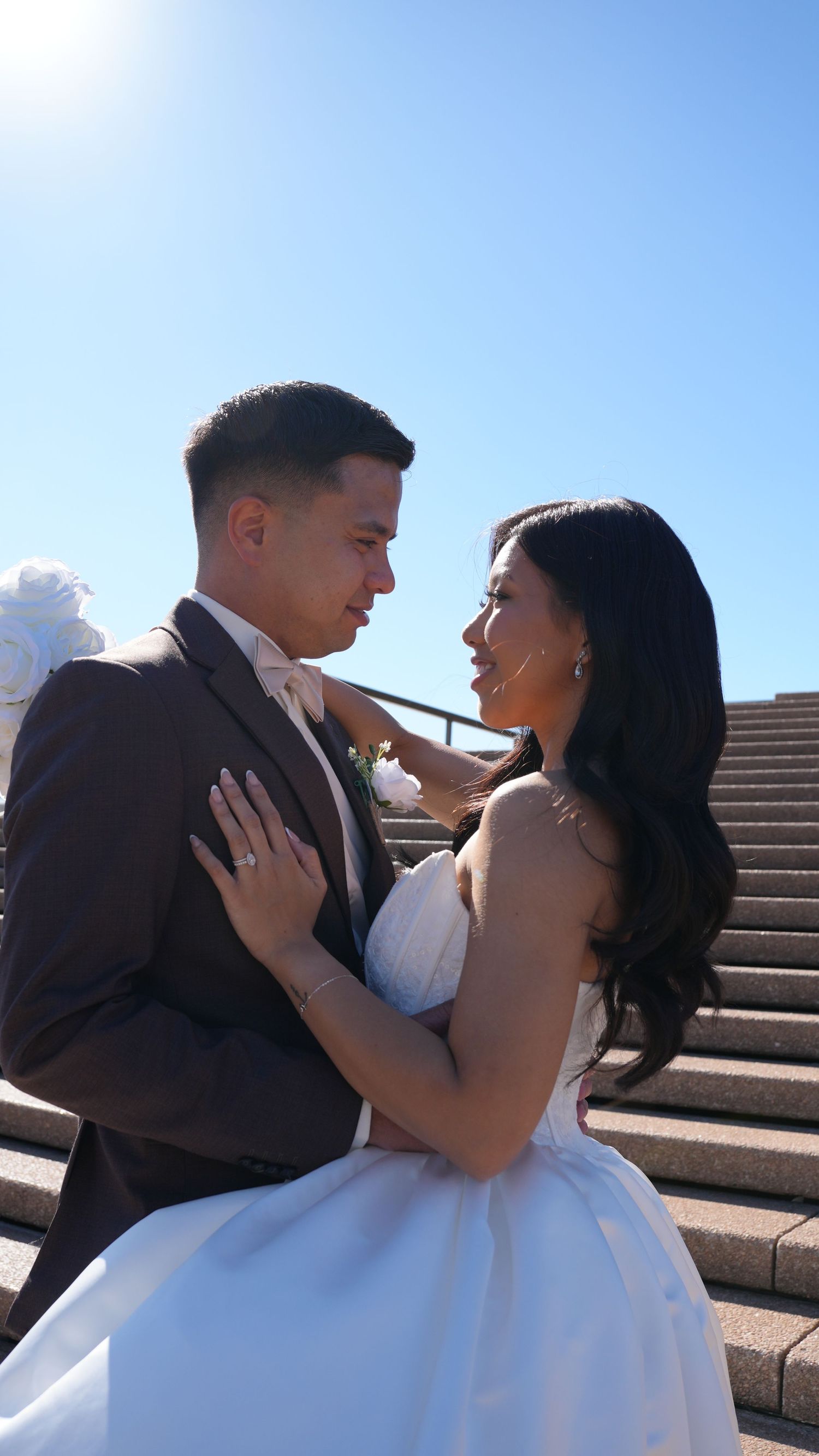 Bride and groom gazing at each other on sunny outdoor steps, she wears white gown, he wears brown suit.