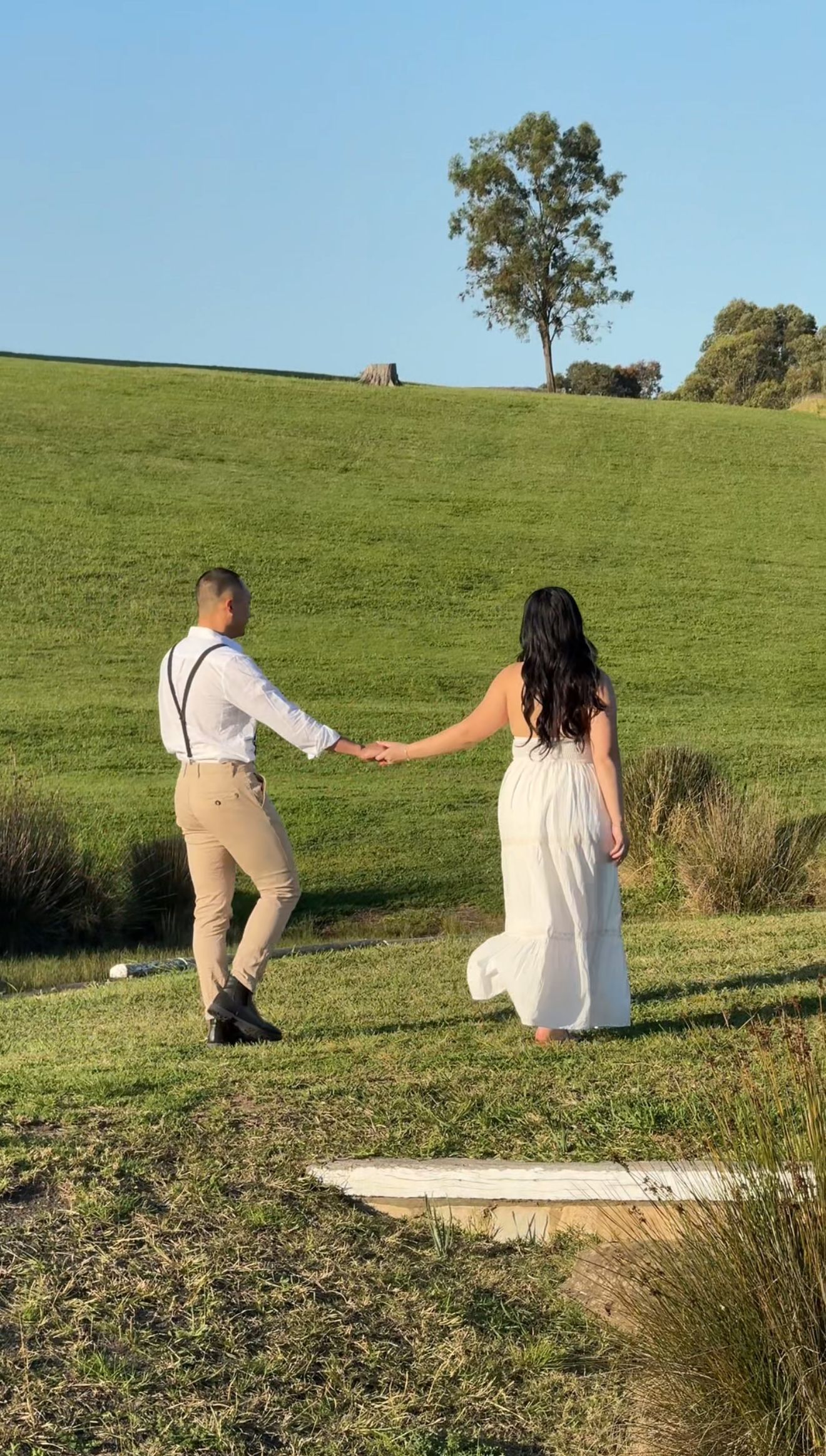 Couple sitting on grass hillside during golden hour with lone tree in background.