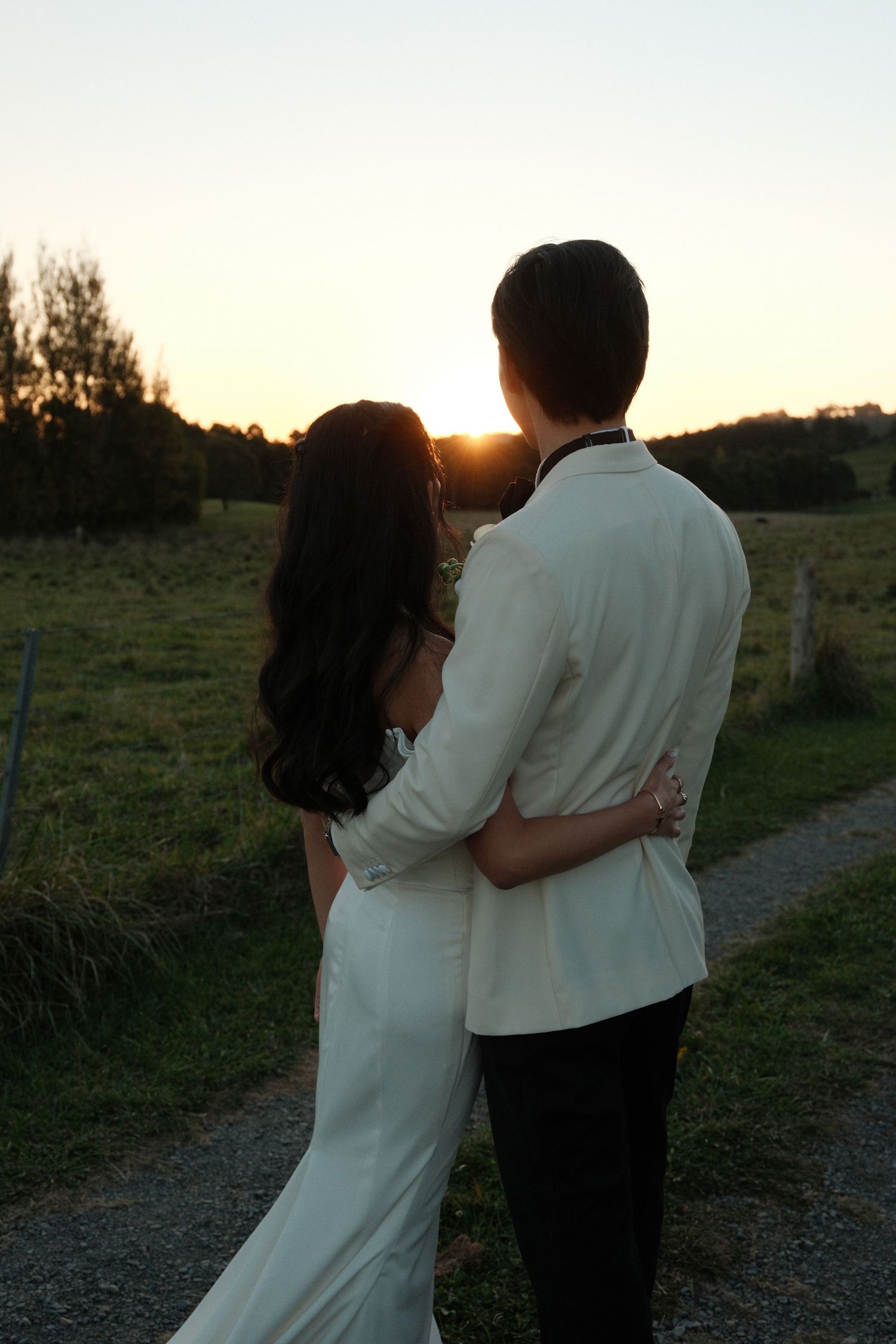 A couple in white wedding attire embraces while watching a golden sunset over a lush green countryside.