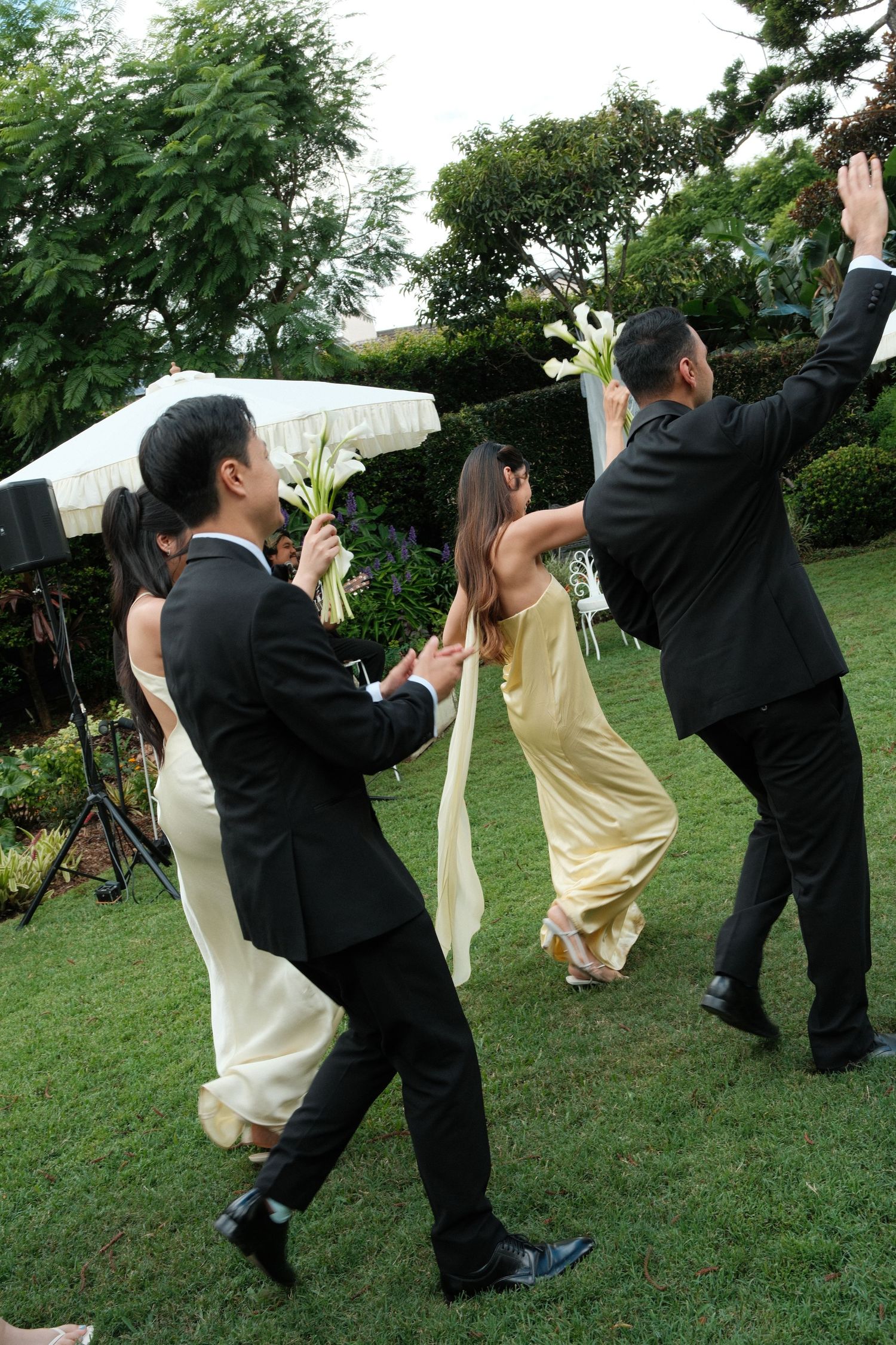Wedding party members in formal attire dance joyfully on a green lawn during an outdoor celebration.