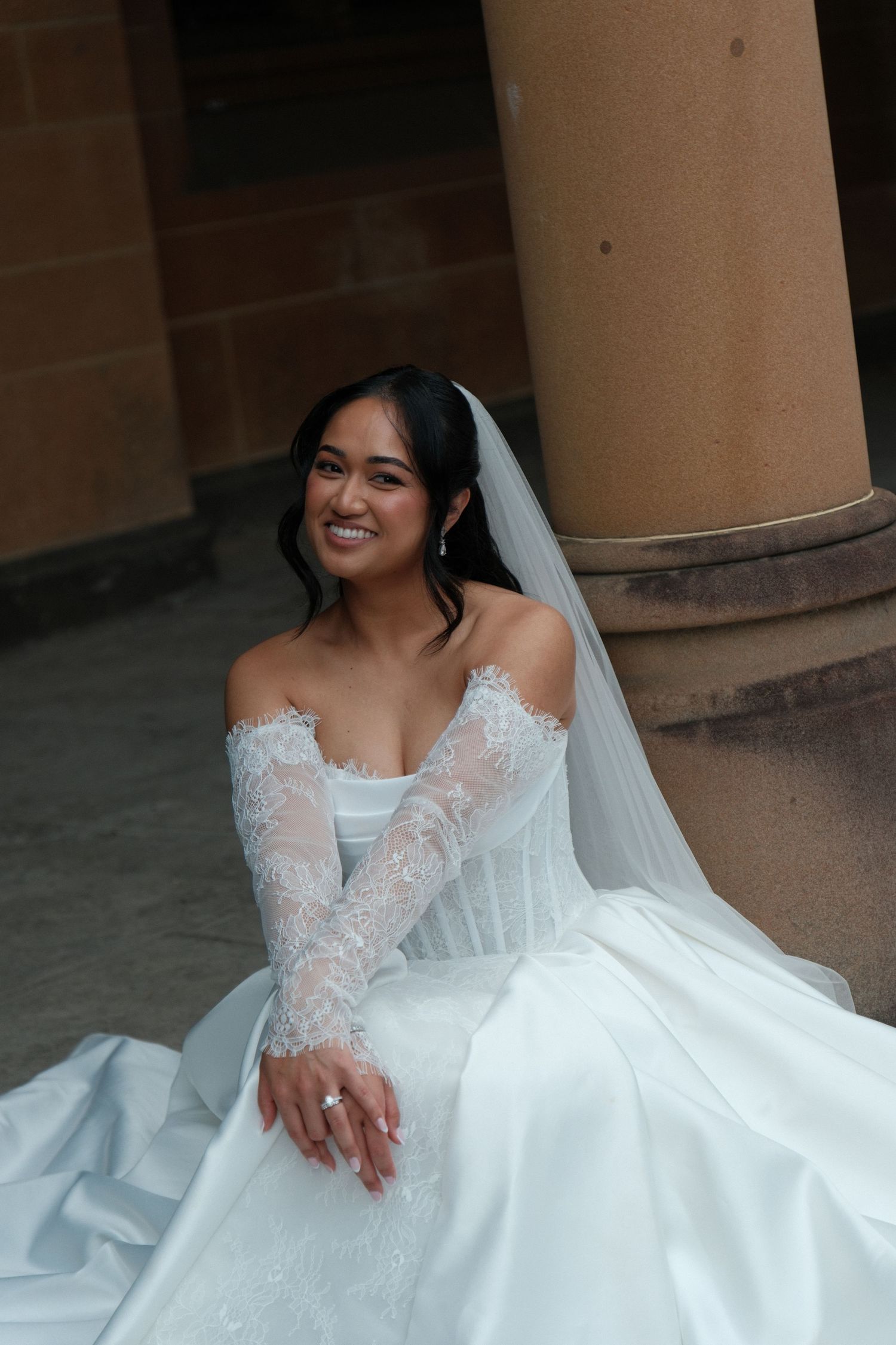 Smiling bride in white lace off-shoulder gown and veil sits gracefully beside stone columns.