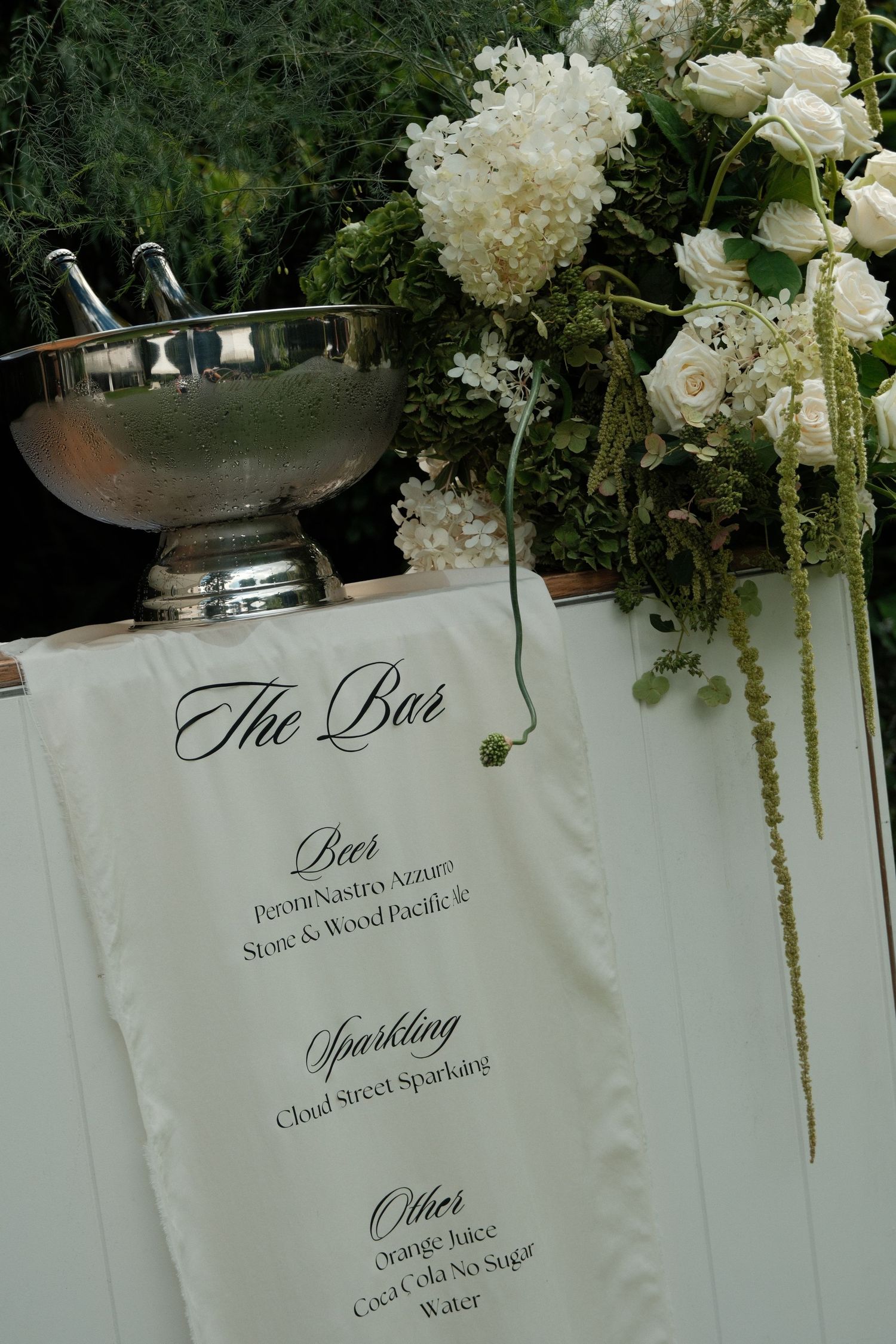 Elegant wedding reception bar menu and floral decorations featuring white hydrangeas and roses in silver vessels.