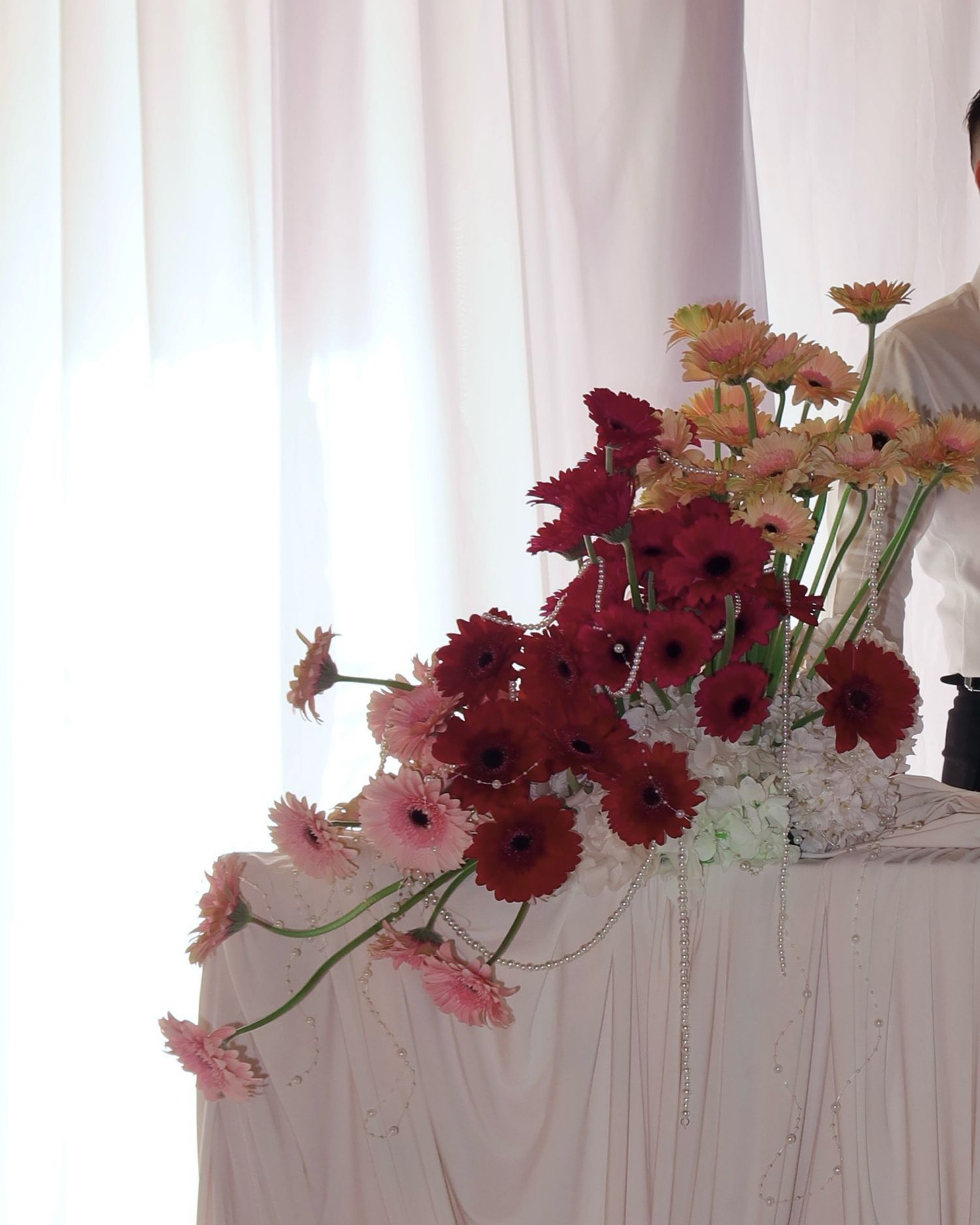 Pink and burgundy flowers arranged in a cascading display along a white mantel or shelf against a light background.