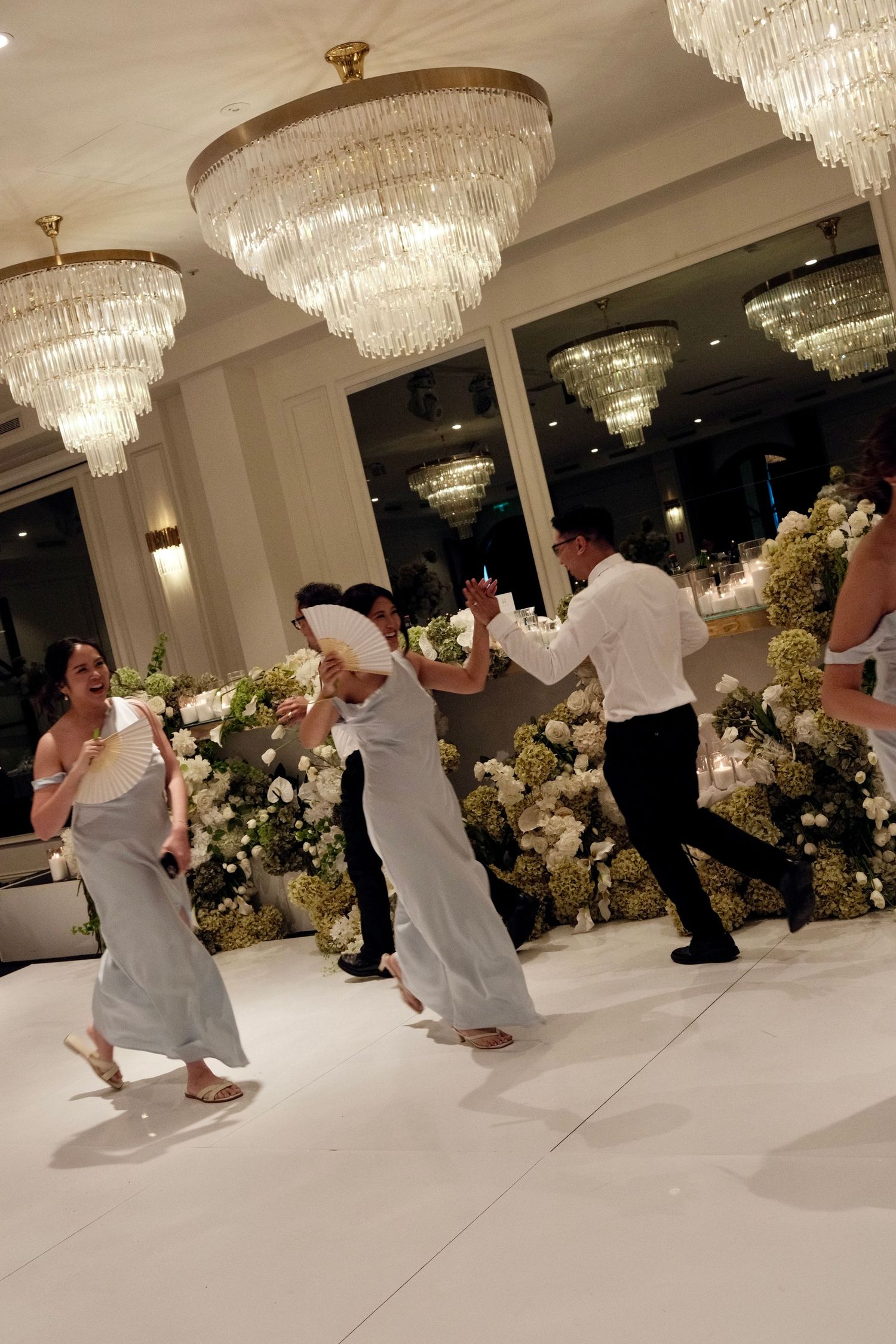 Two people dance together on a white floor under elegant crystal chandeliers at an elegant wedding reception venue.