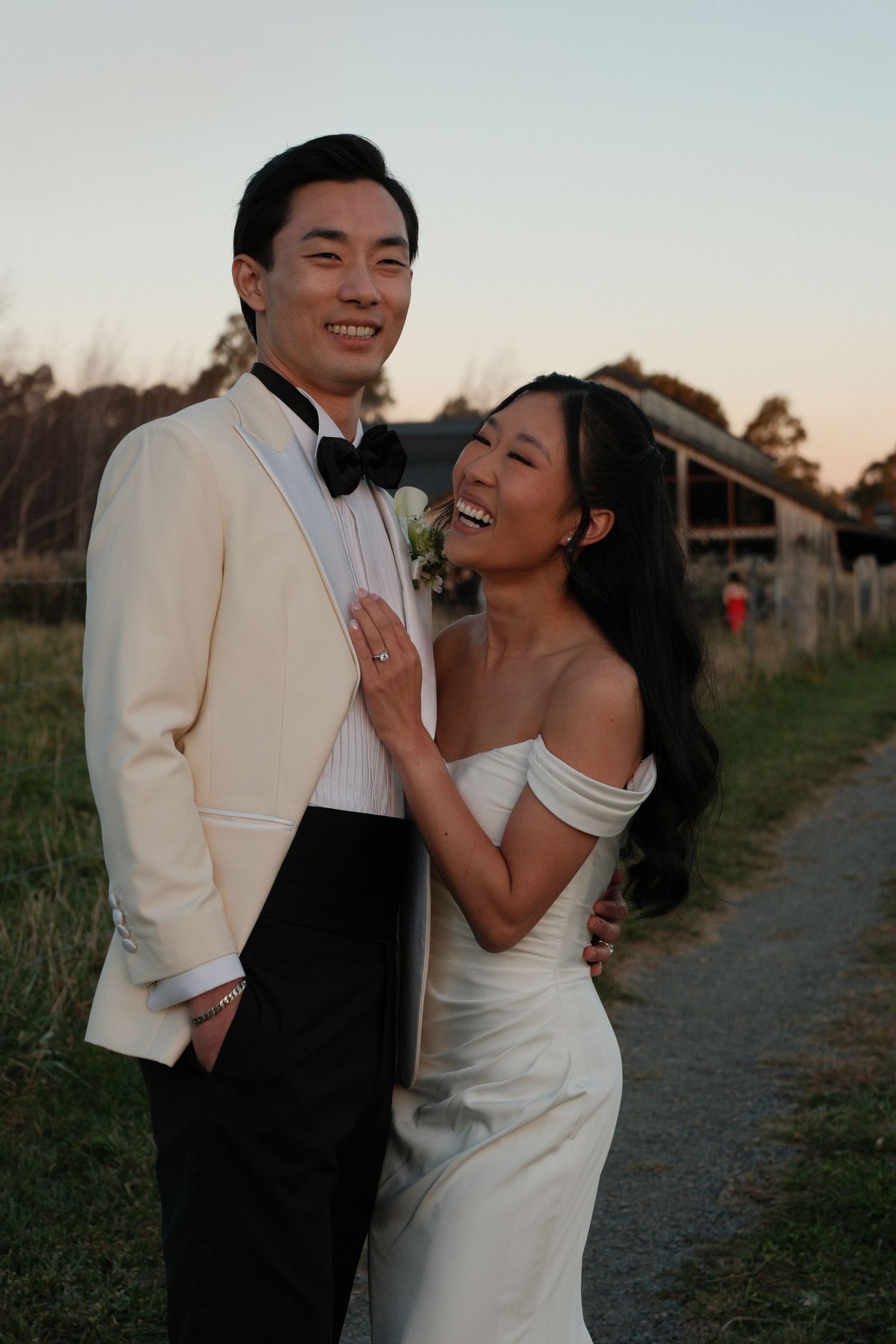 Bride and groom in wedding attire pose at sunset on a rural path, smiling joyfully in elegant formal wear.