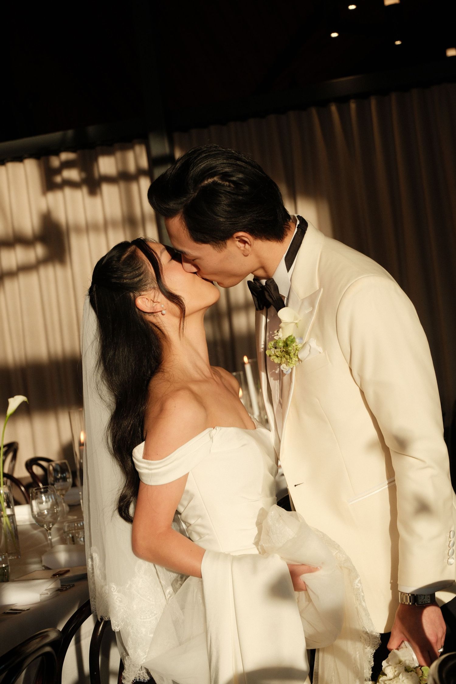 Bride and groom share a kiss at wedding reception, bride in off-shoulder gown, groom in ivory tuxedo.