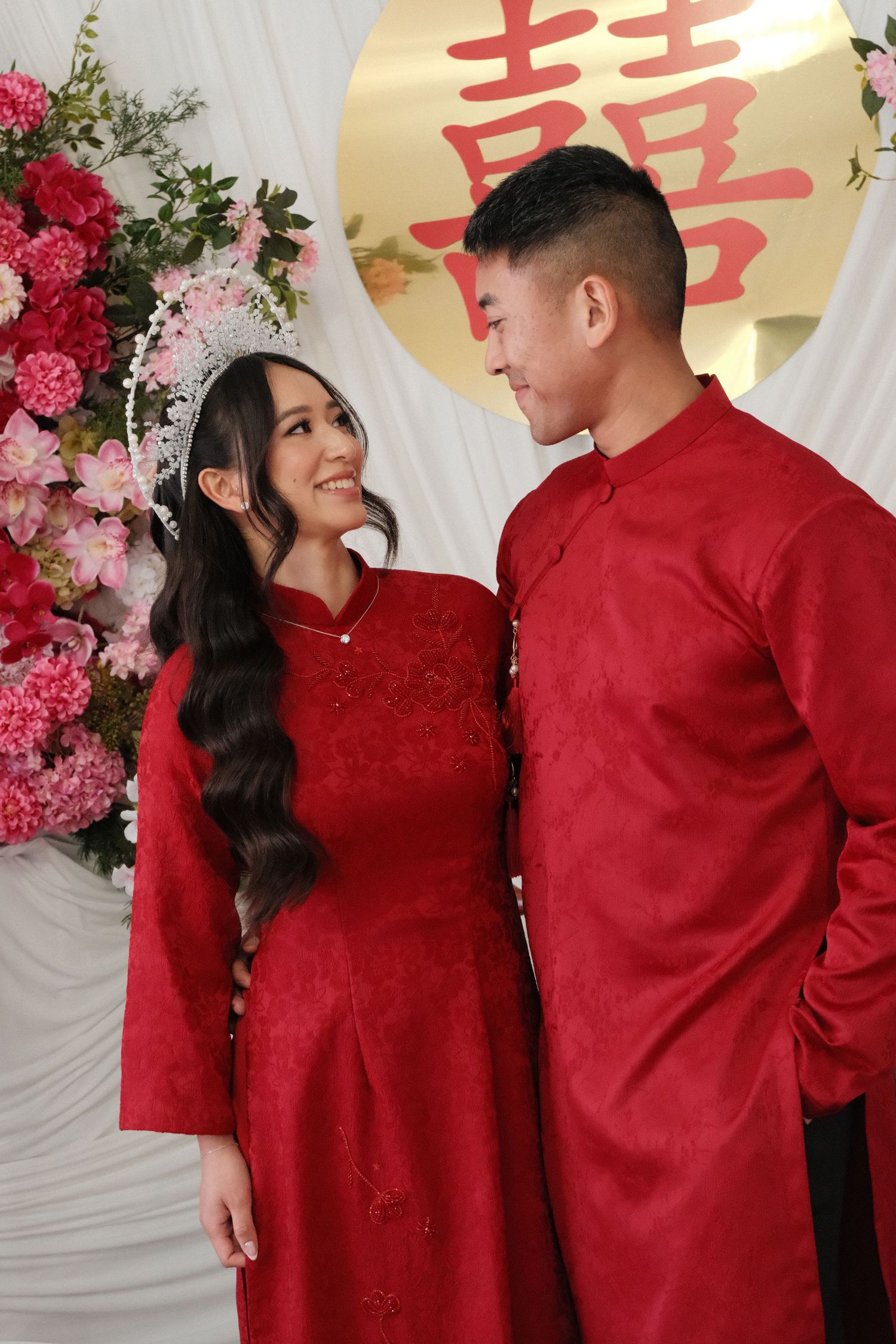 Vietnamese couple in traditional red ao dai attire posing together at a wedding ceremony with floral decorations.