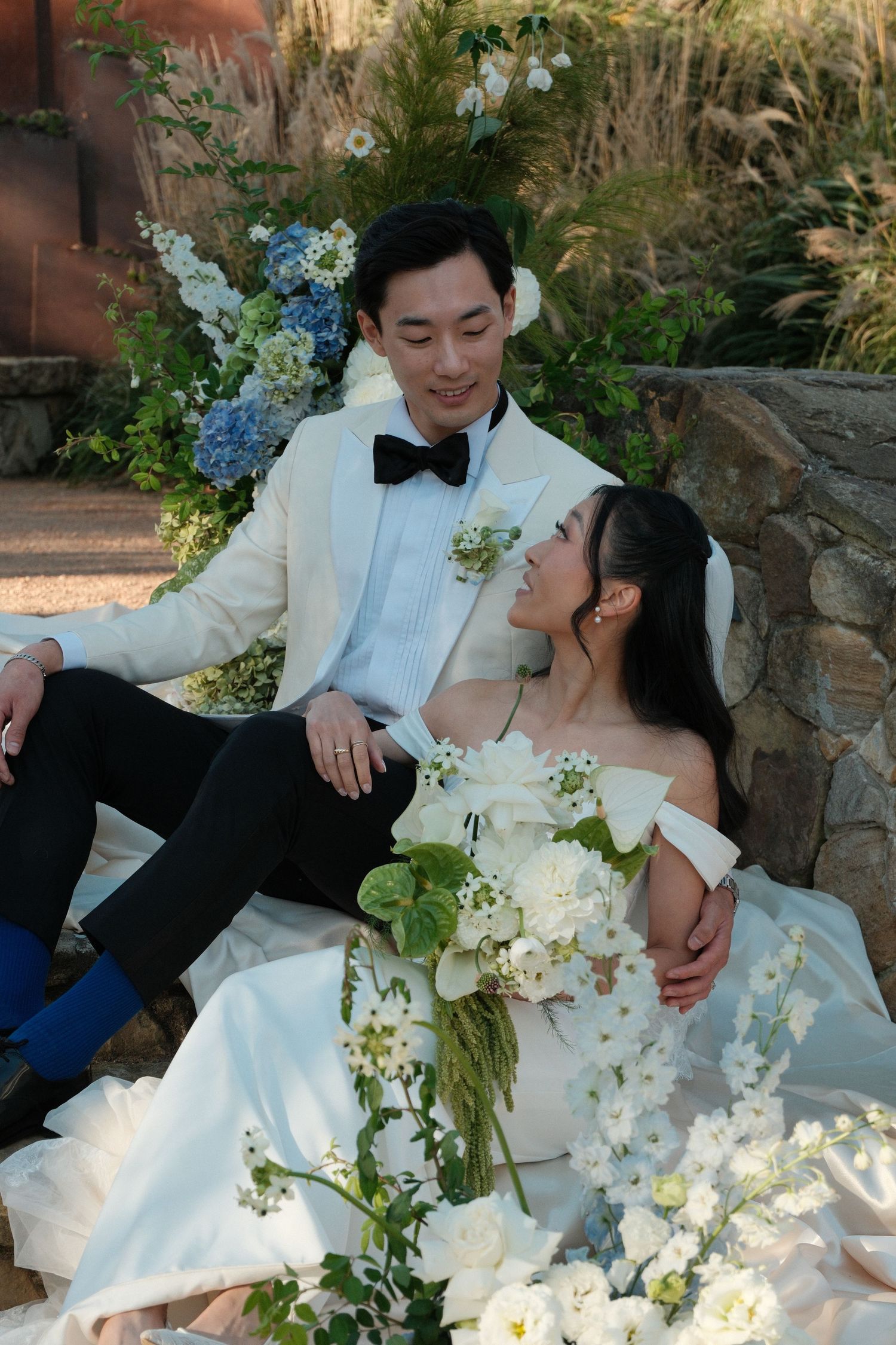 Bride and groom in elegant wedding attire sitting together among white and blue floral arrangements outdoors.