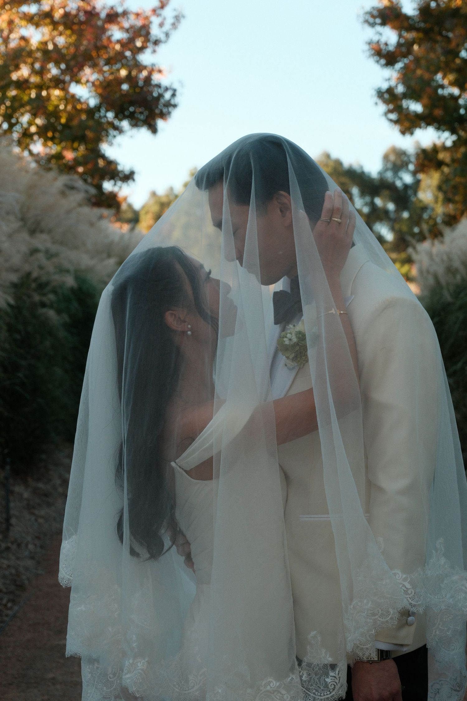 Bride and groom sharing an intimate moment under a white lace wedding veil outdoors surrounded by autumn trees.
