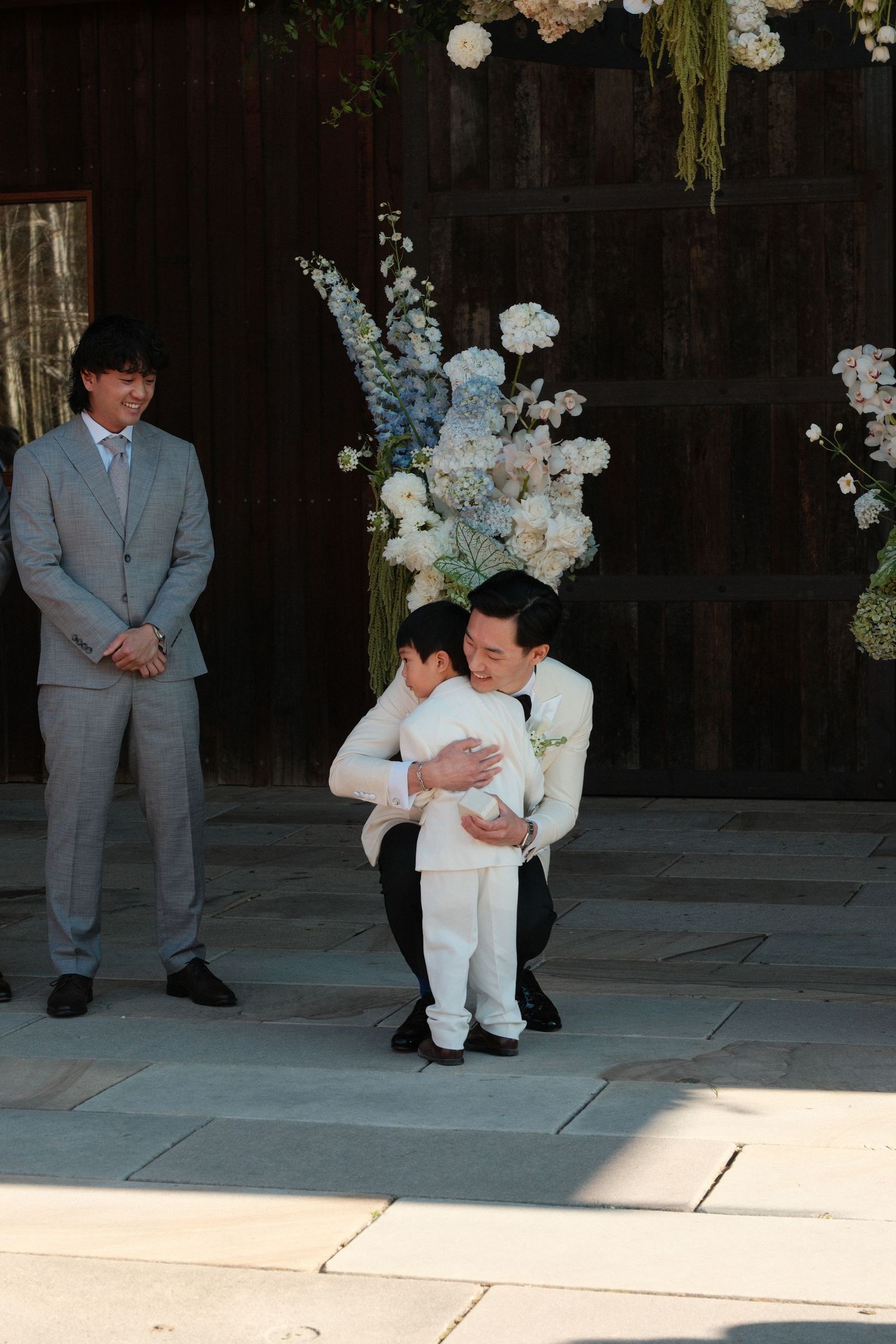 A groom in a white suit crouches to hug a young child at an elegant wedding ceremony with floral arrangements.