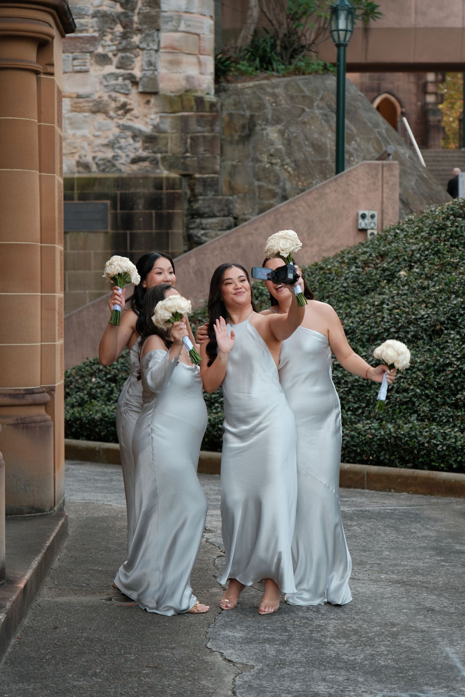 Three bridesmaids in silver satin gowns holding white bouquets pose playfully outside a stone building.