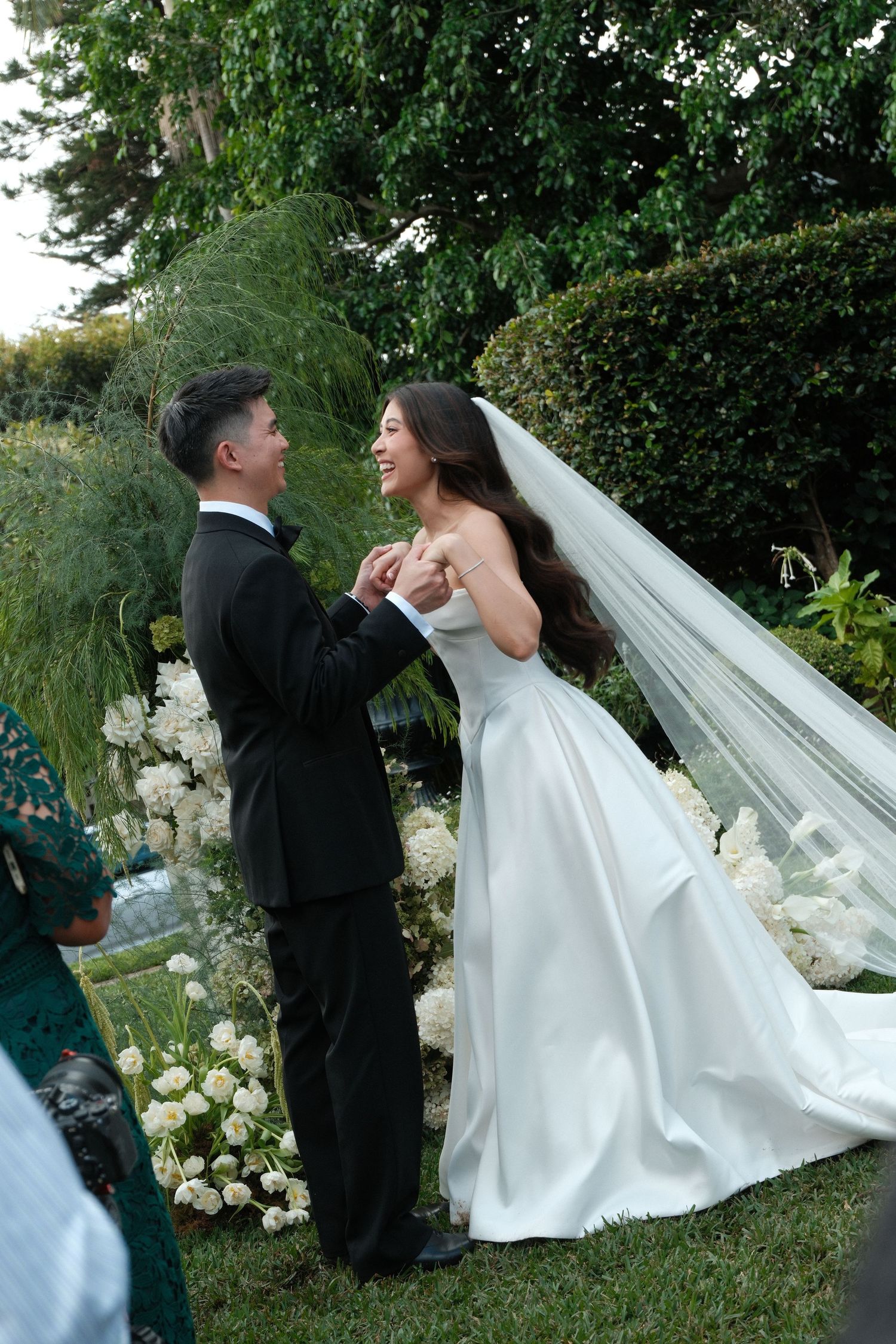 A romantic wedding couple shares a playful moment together in a lush garden setting with white floral decorations.