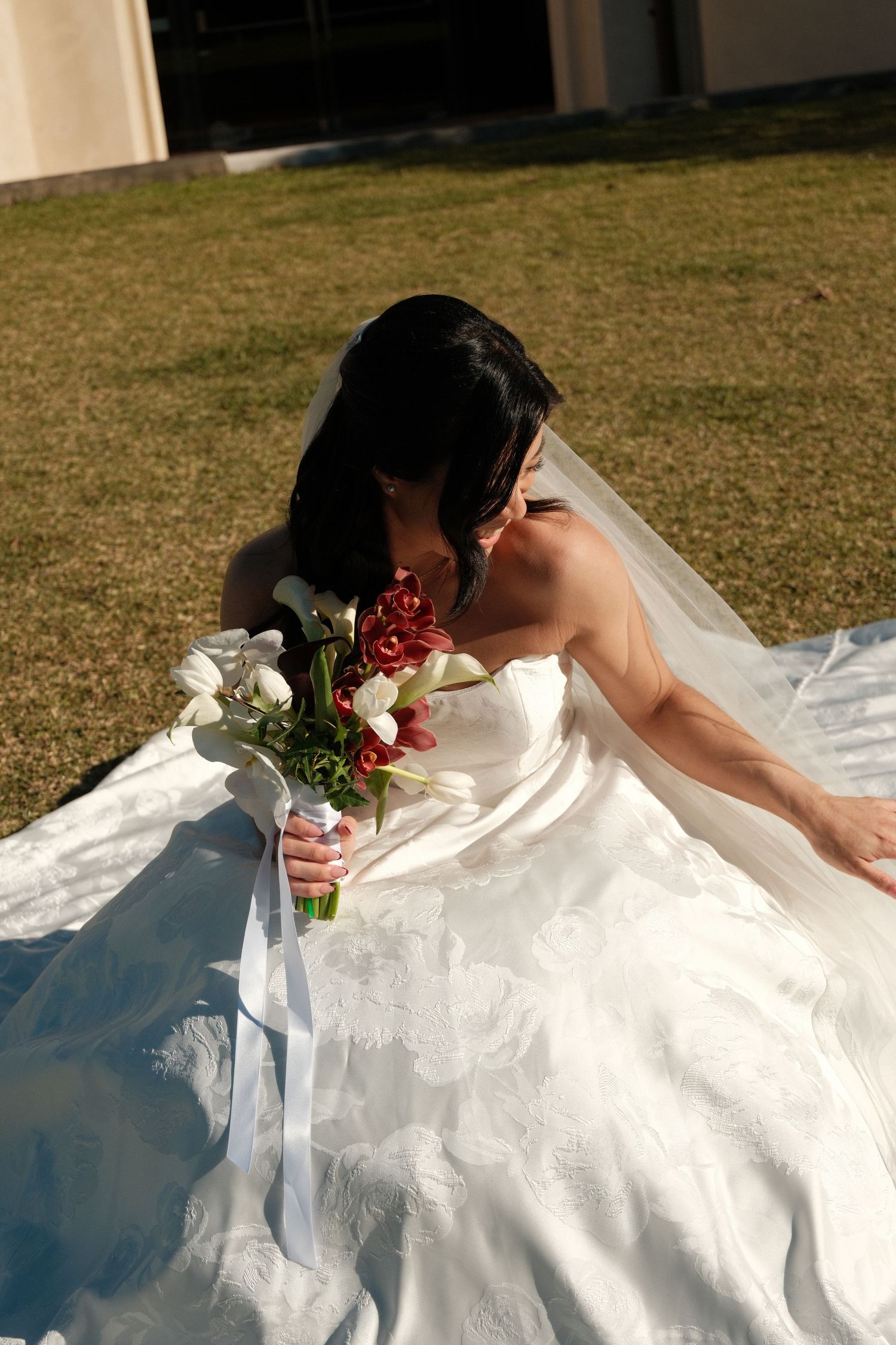 Bride in white strapless ball gown sits on grass holding red and white floral bouquet with flowing veil on sunny day.