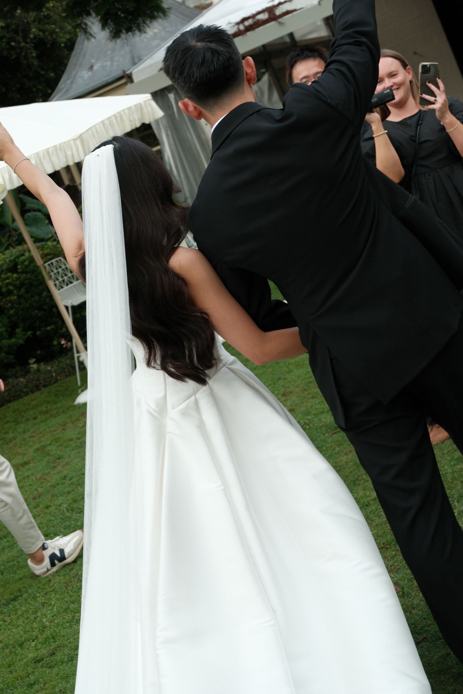 A couple in wedding attire dips dramatically during their first dance on a grassy lawn while guests look on.