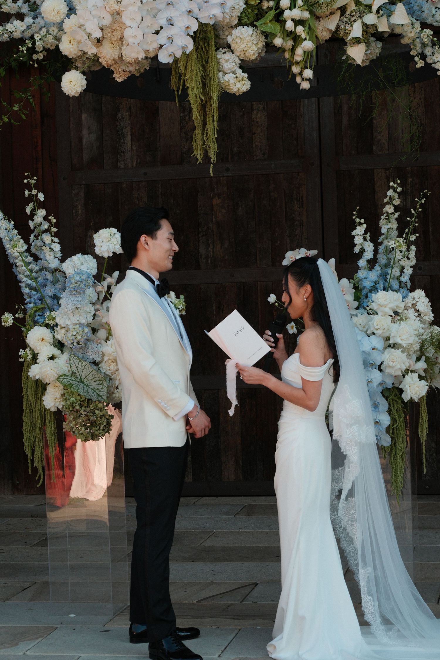Couple exchanging vows at floral arch wedding ceremony with white and blue flowers, bride in white gown with veil.