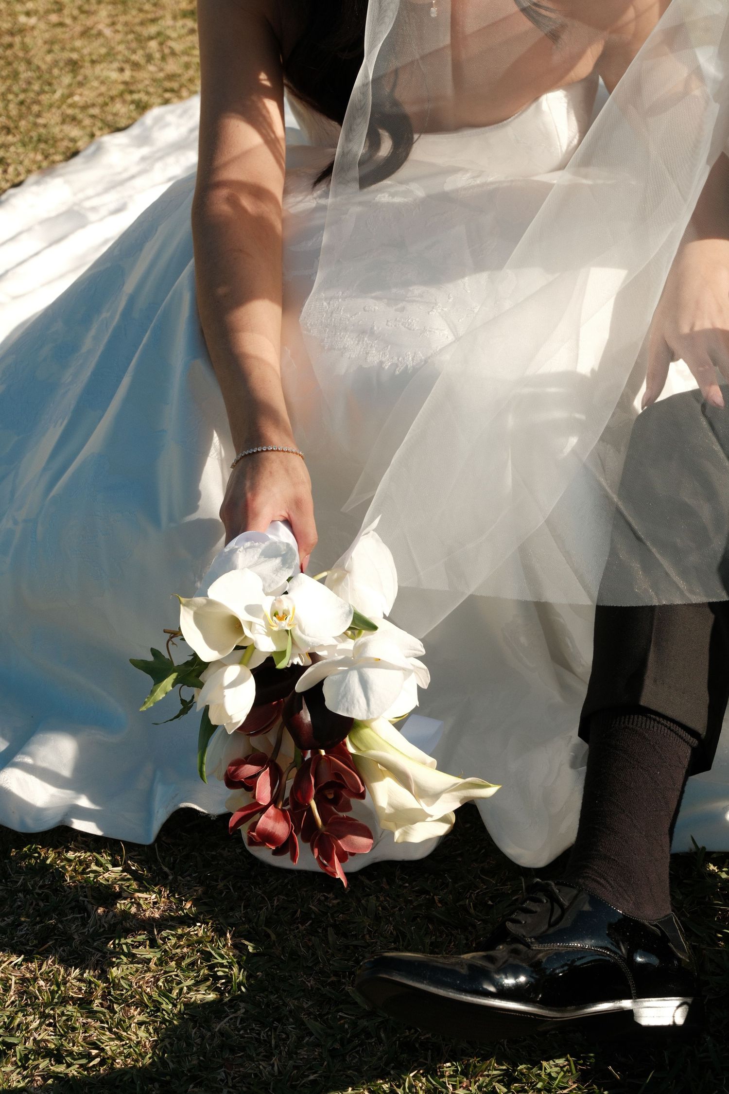 Bride in white gown holding elegant bouquet of white orchids and calla lilies beside groom in black shoes on grass.