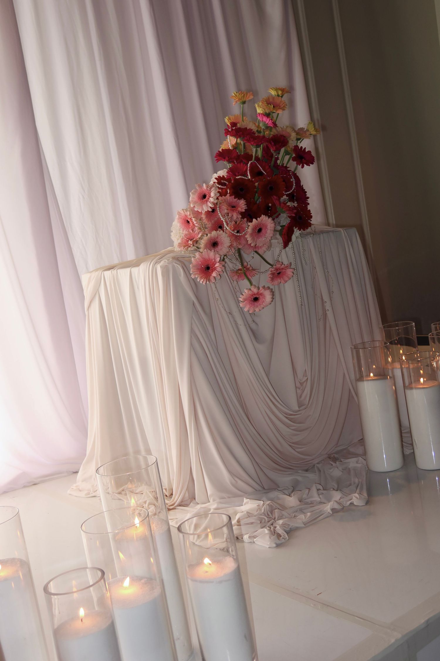 White draped fabric backdrop with glowing candles and pink floral arrangements creates an elegant wedding ceremony setting.