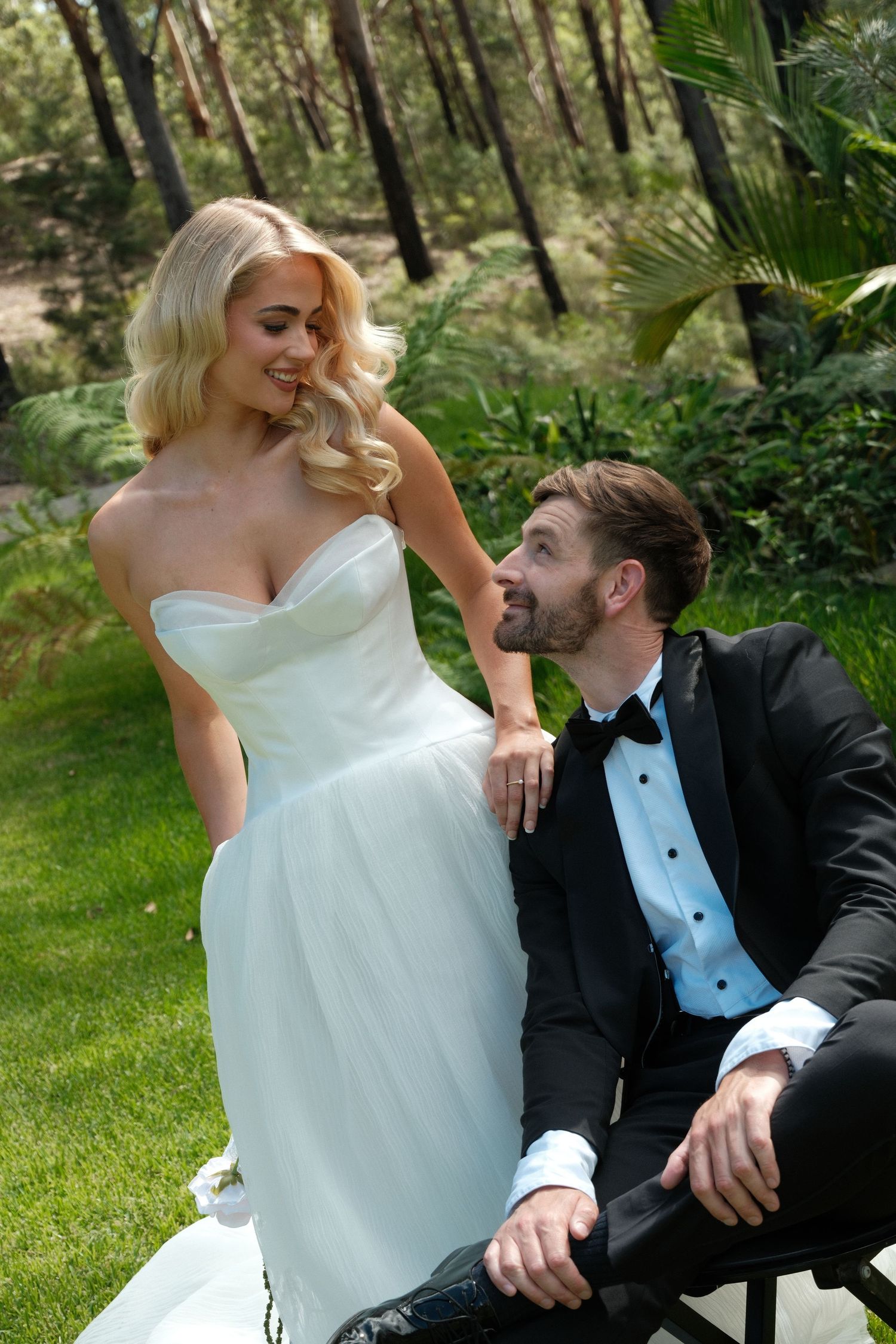 A couple in wedding attire shares a joyful moment together while sitting on grass surrounded by lush greenery.