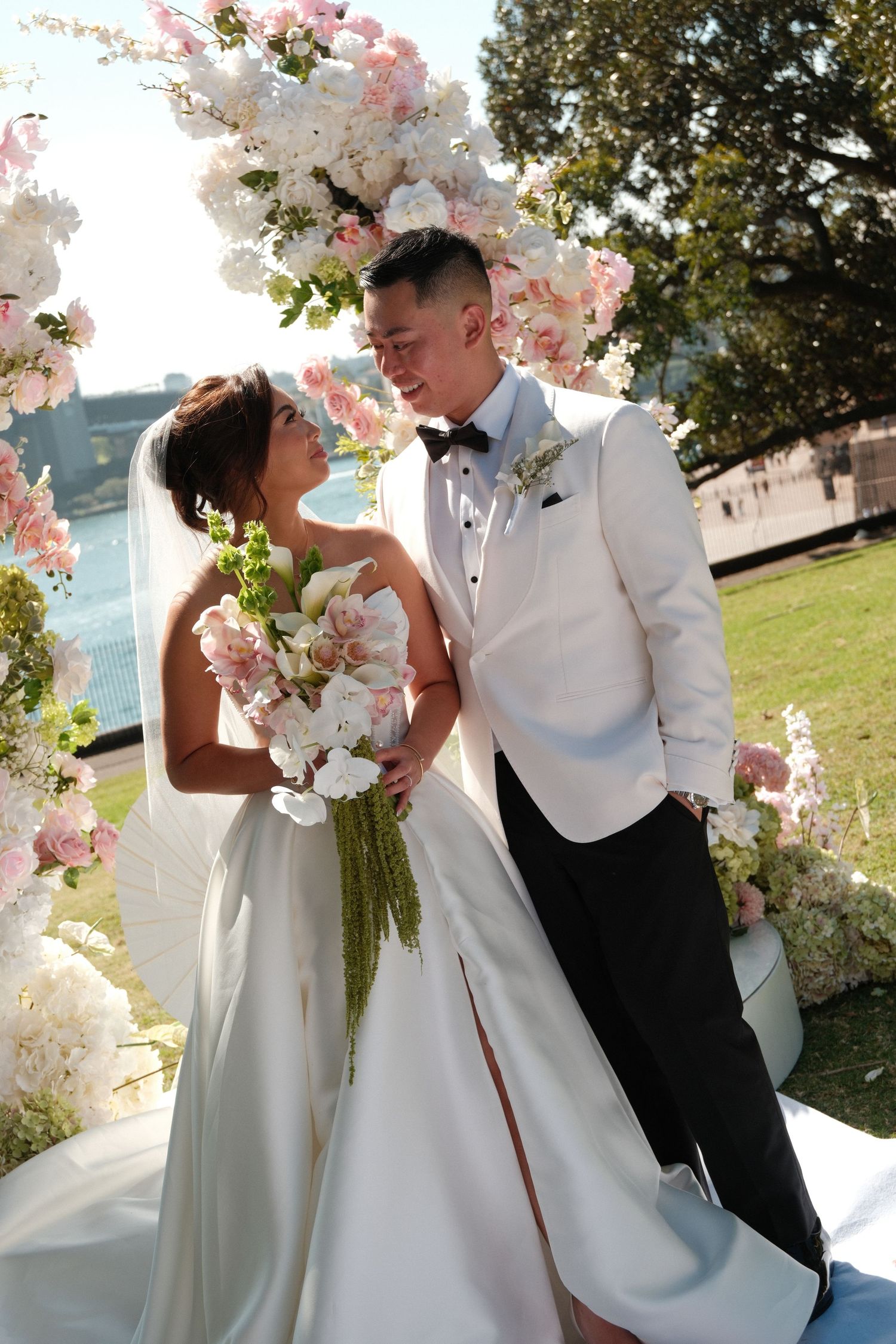 Bride and groom share a romantic moment at outdoor wedding ceremony surrounded by pink and white floral arch near waterfront.