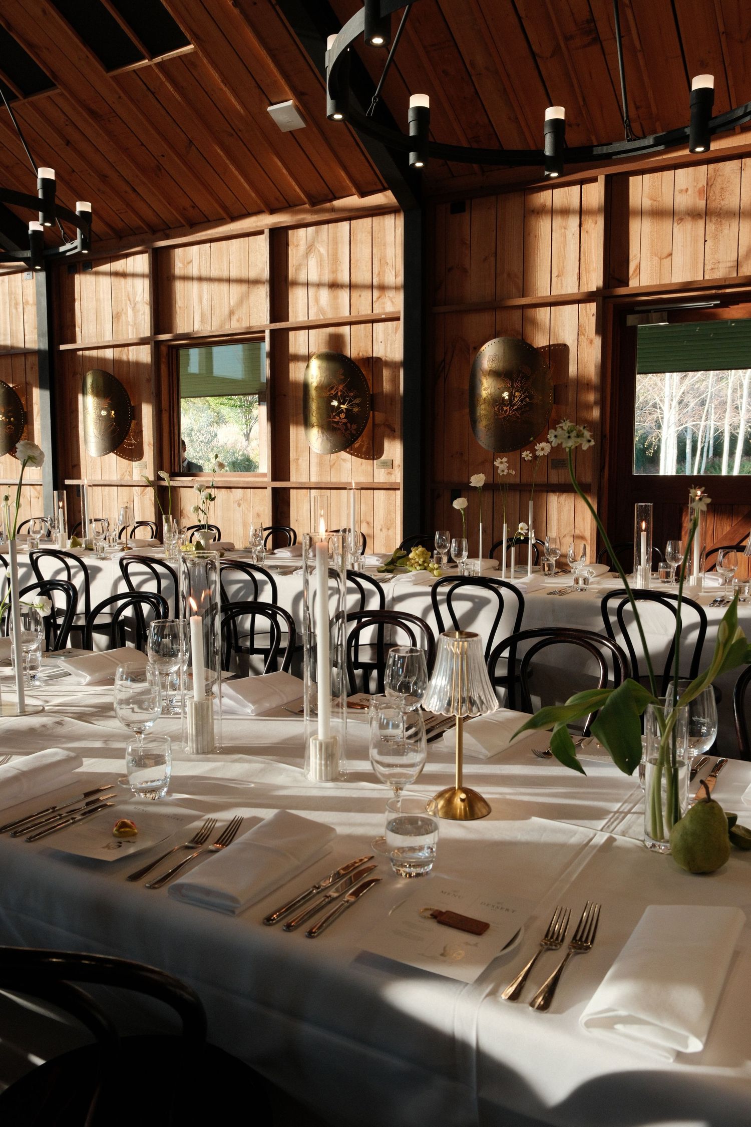 Elegant wedding reception venue with long white-clothed tables, black bentwood chairs, gold accents, and warm wood-paneled walls.