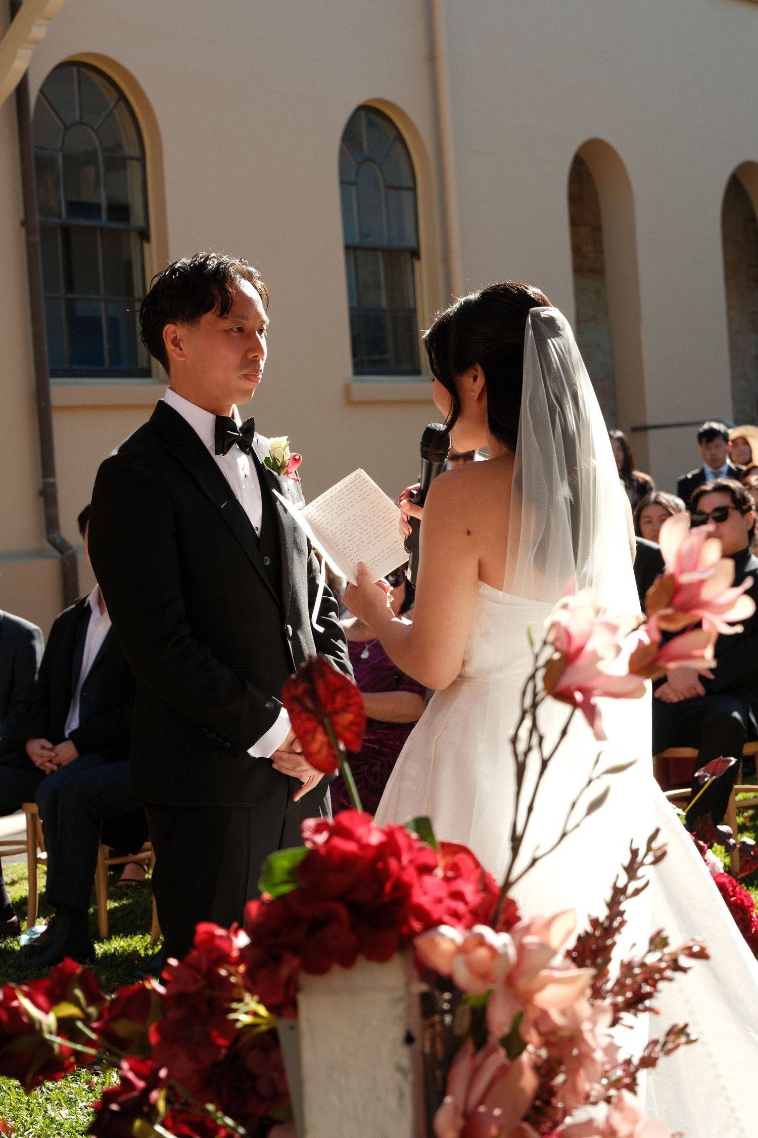 Couple exchanging vows at outdoor wedding ceremony with red and pink floral arrangements near a church.