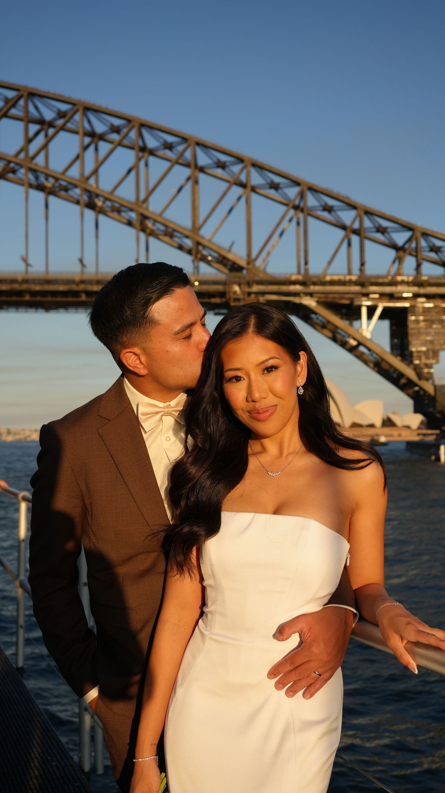 Couple in wedding attire kissing near Sydney Harbour Bridge and Opera House at golden hour.