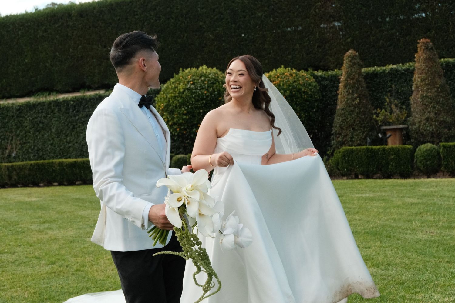 Couple in wedding attire smiling in a garden, bride in white strapless gown, groom in white suit with bow tie.