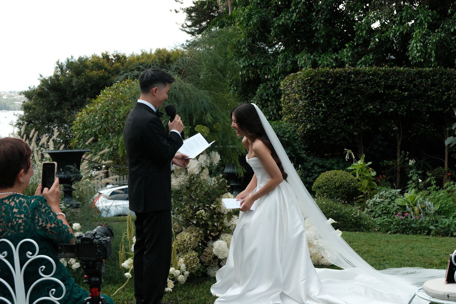 A wedding ceremony takes place in a lush garden setting with white decor and floral arrangements.
