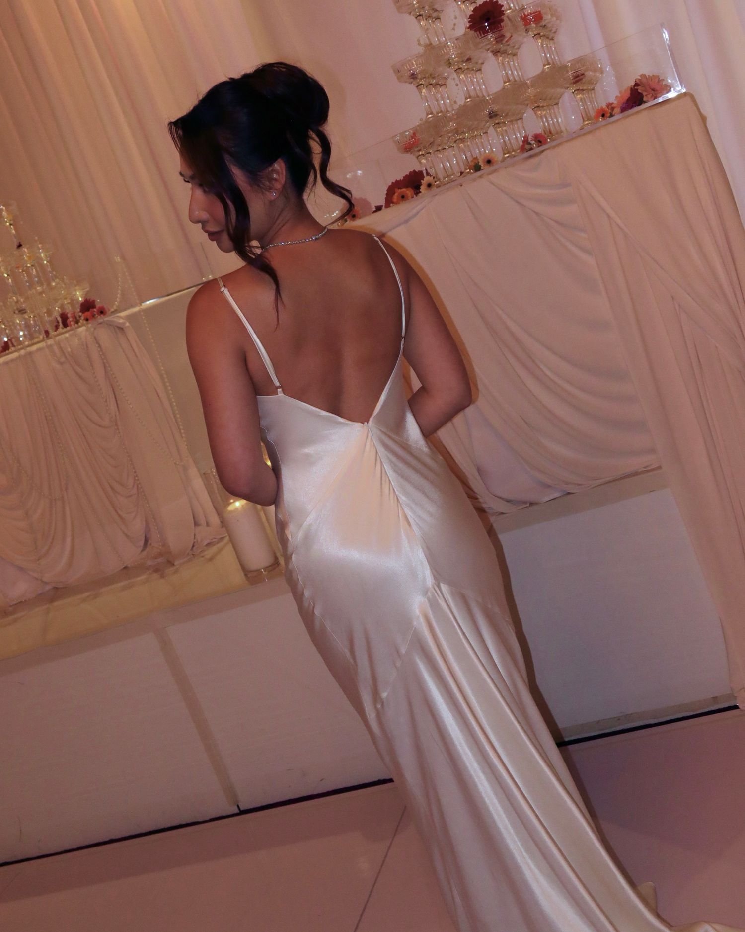 A person wearing an elegant white silk gown poses from behind in a decorated event space.