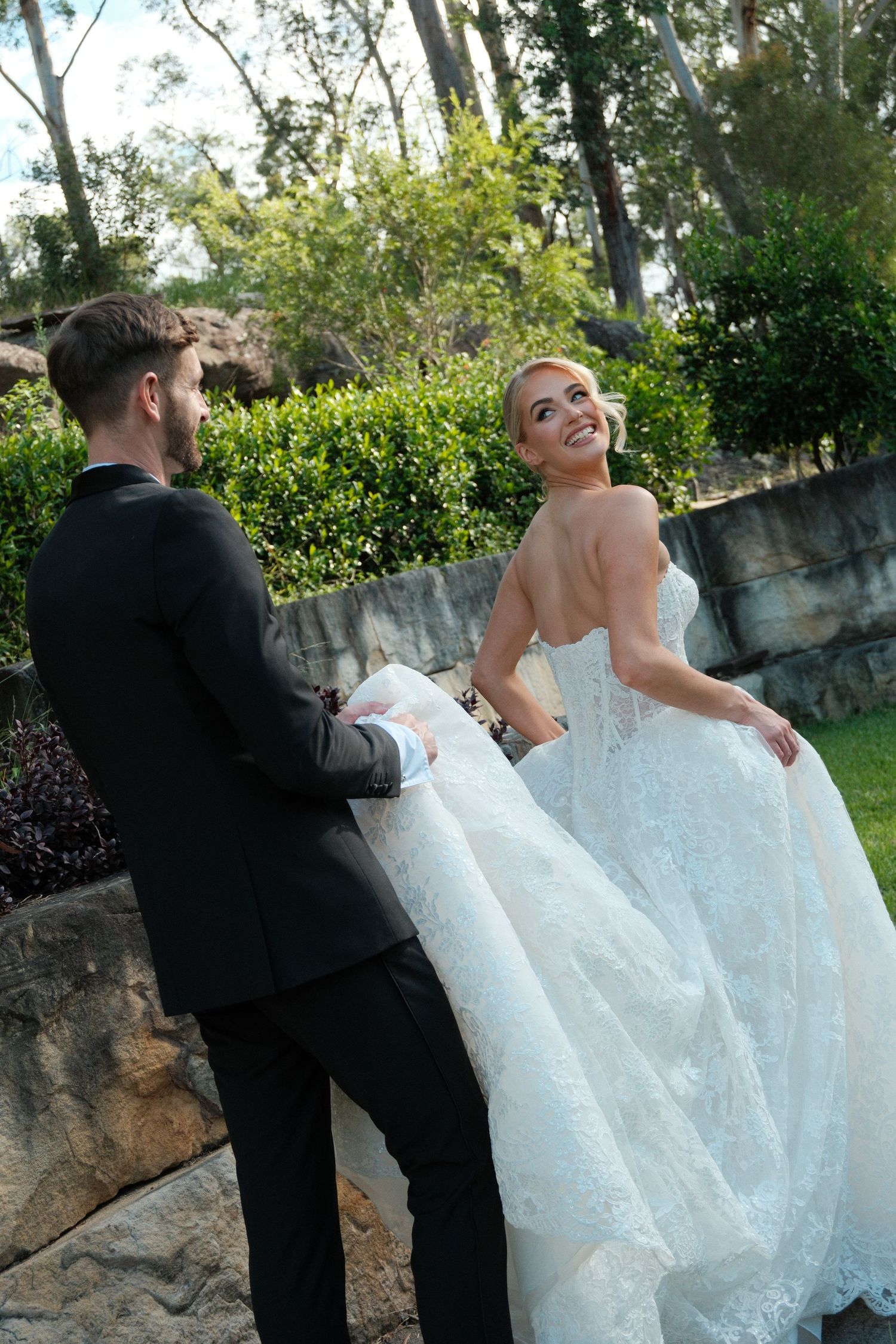 A couple in formal wedding attire share playful moments together in a garden setting with stone walls and greenery.
