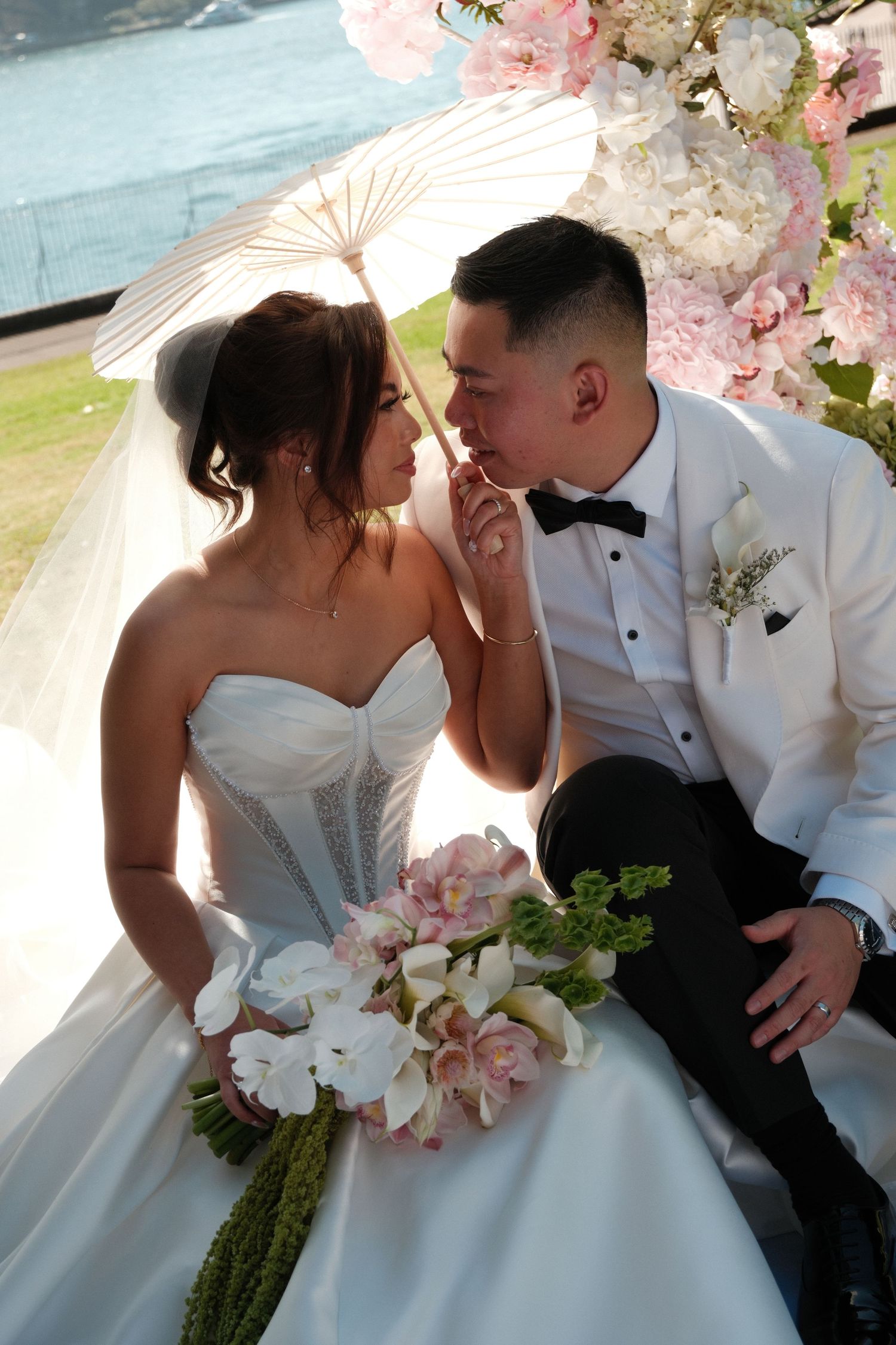Asian bride and groom share romantic moment under white parasol, surrounded by pink floral arch near waterfront.
