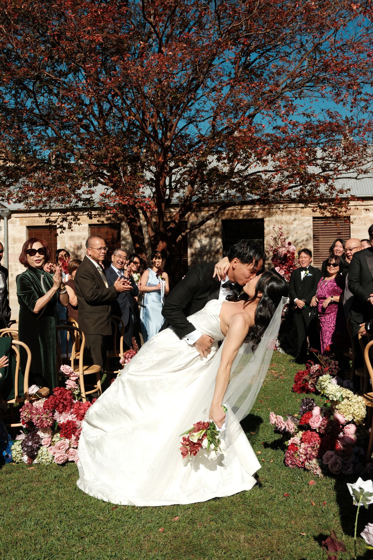 Bride and groom share a romantic dip kiss at outdoor wedding ceremony with guests applauding beside floral arrangements.