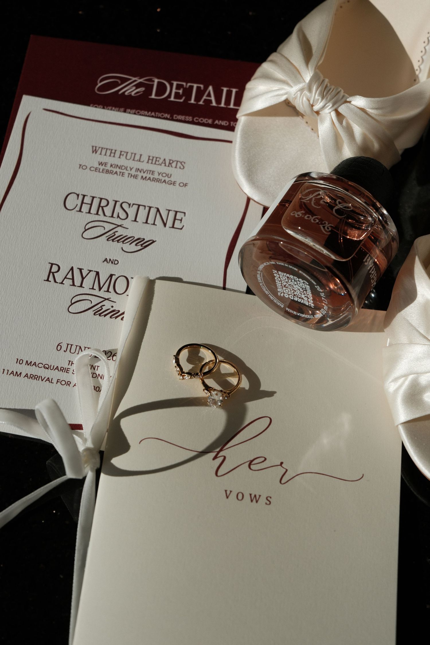 Wedding flat lay featuring vow books, gold rings, perfume bottle, and marriage invitation for Christine and Raymond.