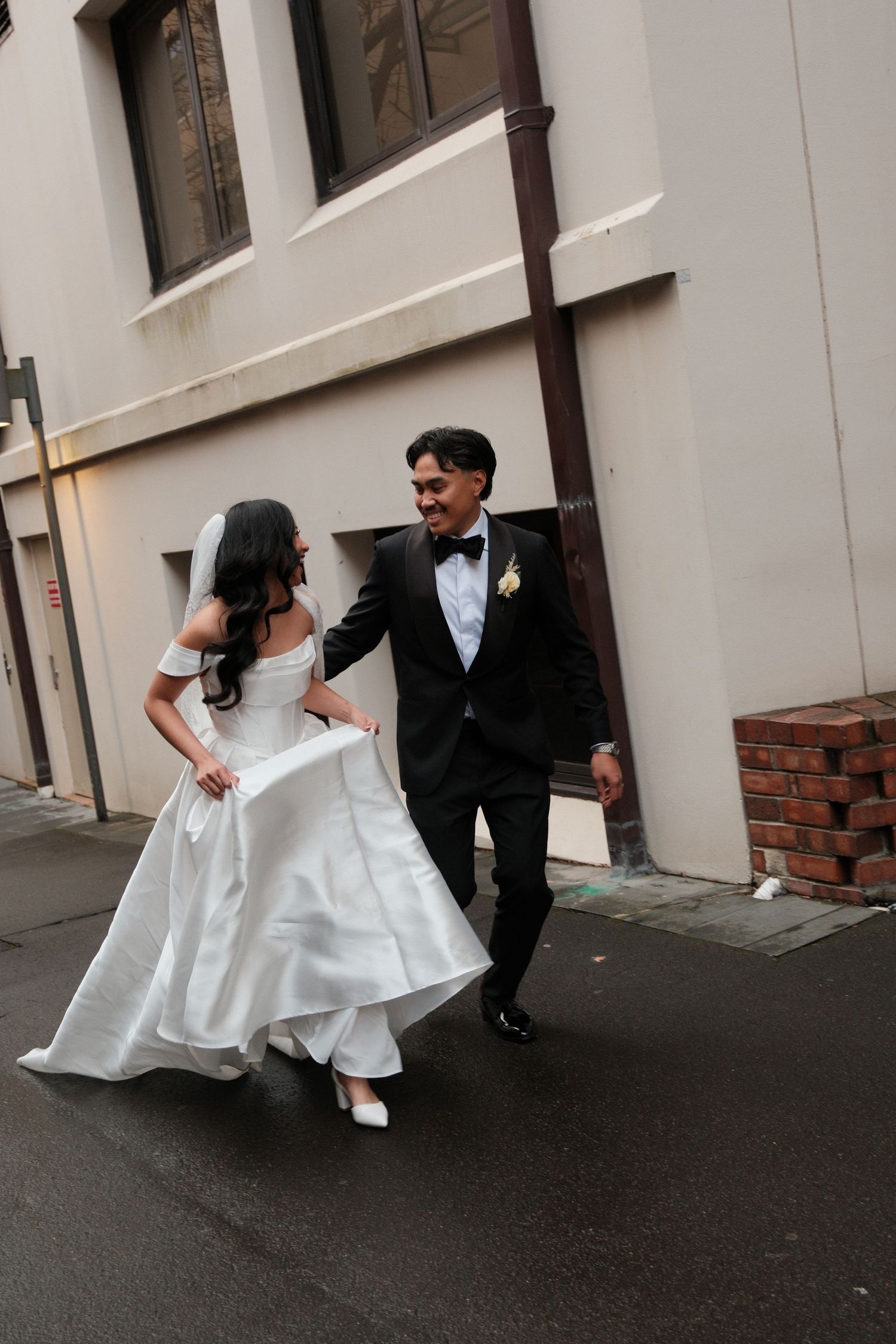 A joyful bride in a white off-shoulder gown and groom in black tuxedo walk together down a city alley.