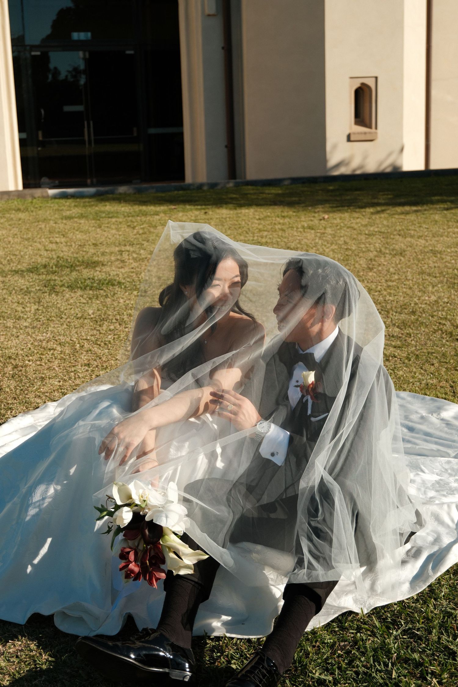 Bride and groom sit together on grass, sharing an intimate moment under the bride's flowing wedding veil.