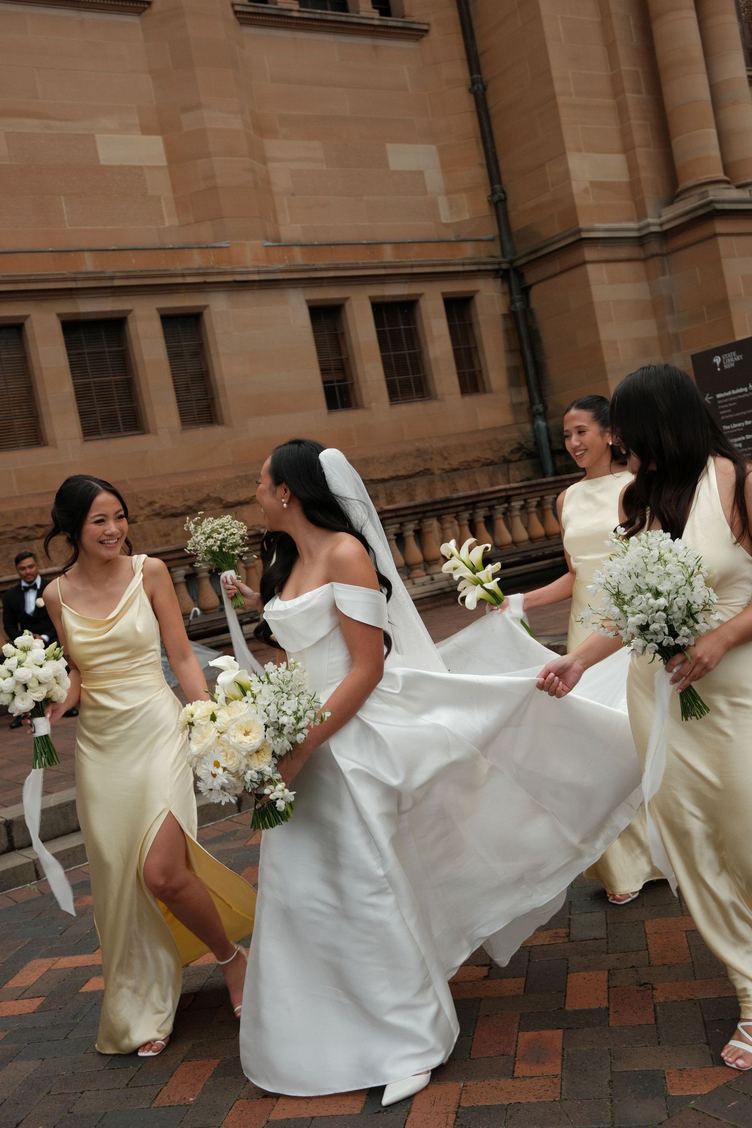 Bride in white gown with bridesmaids in gold dresses holding white bouquets outside a stone church.