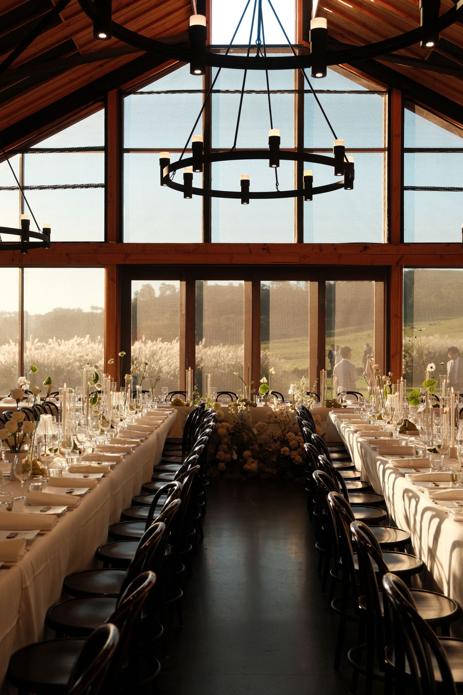 Elegant wedding reception hall with long white tables, floral centerpieces, black chairs, chandelier, and panoramic vineyard views.