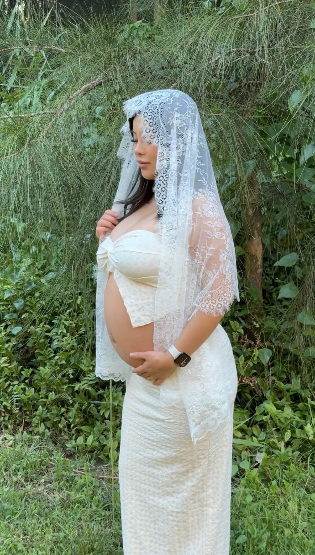 A pregnant figure in a white lace dress and veil poses outdoors against a green natural backdrop.