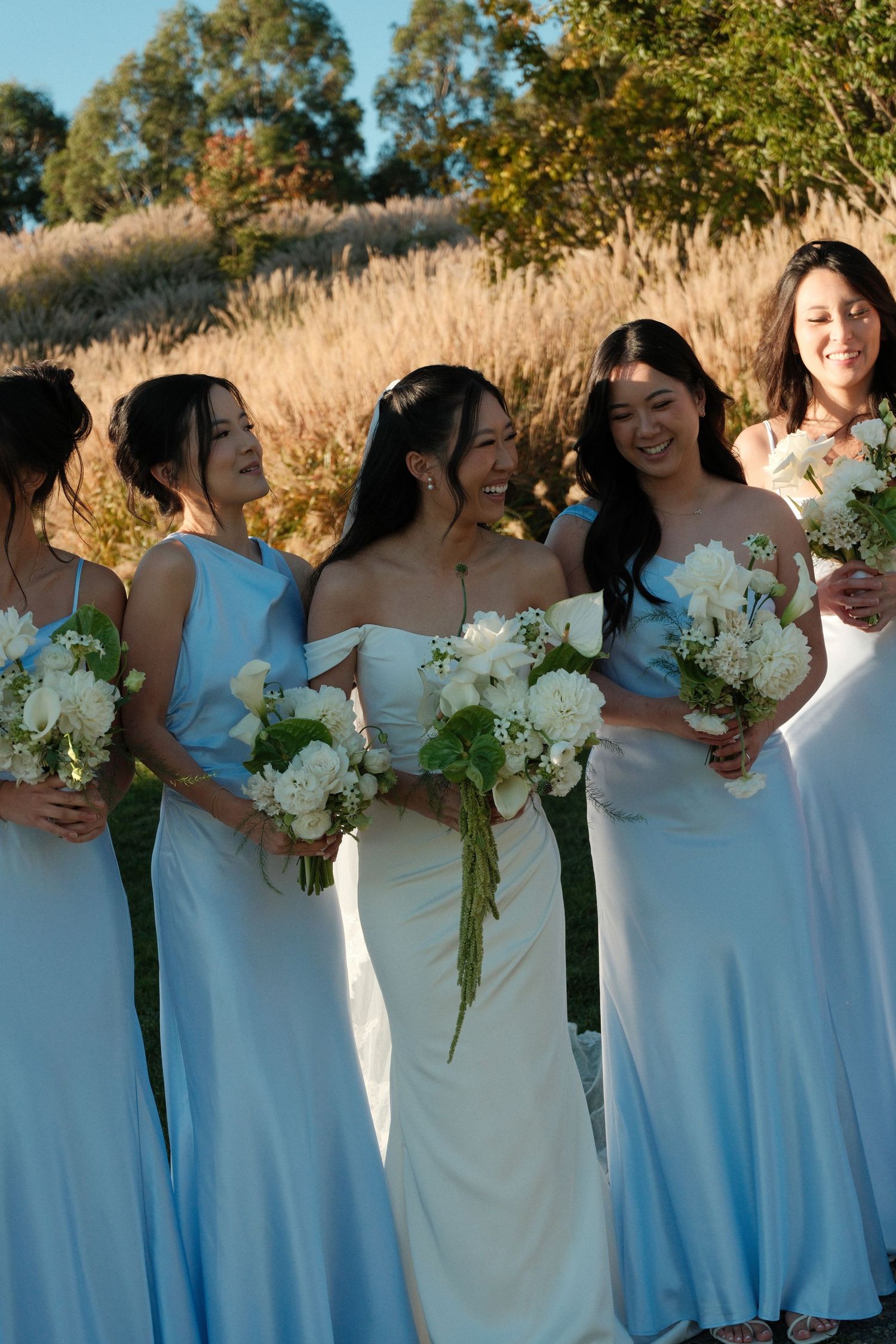 Bride with bridesmaids in light blue gowns holding white bouquets outdoors at golden hour wedding.