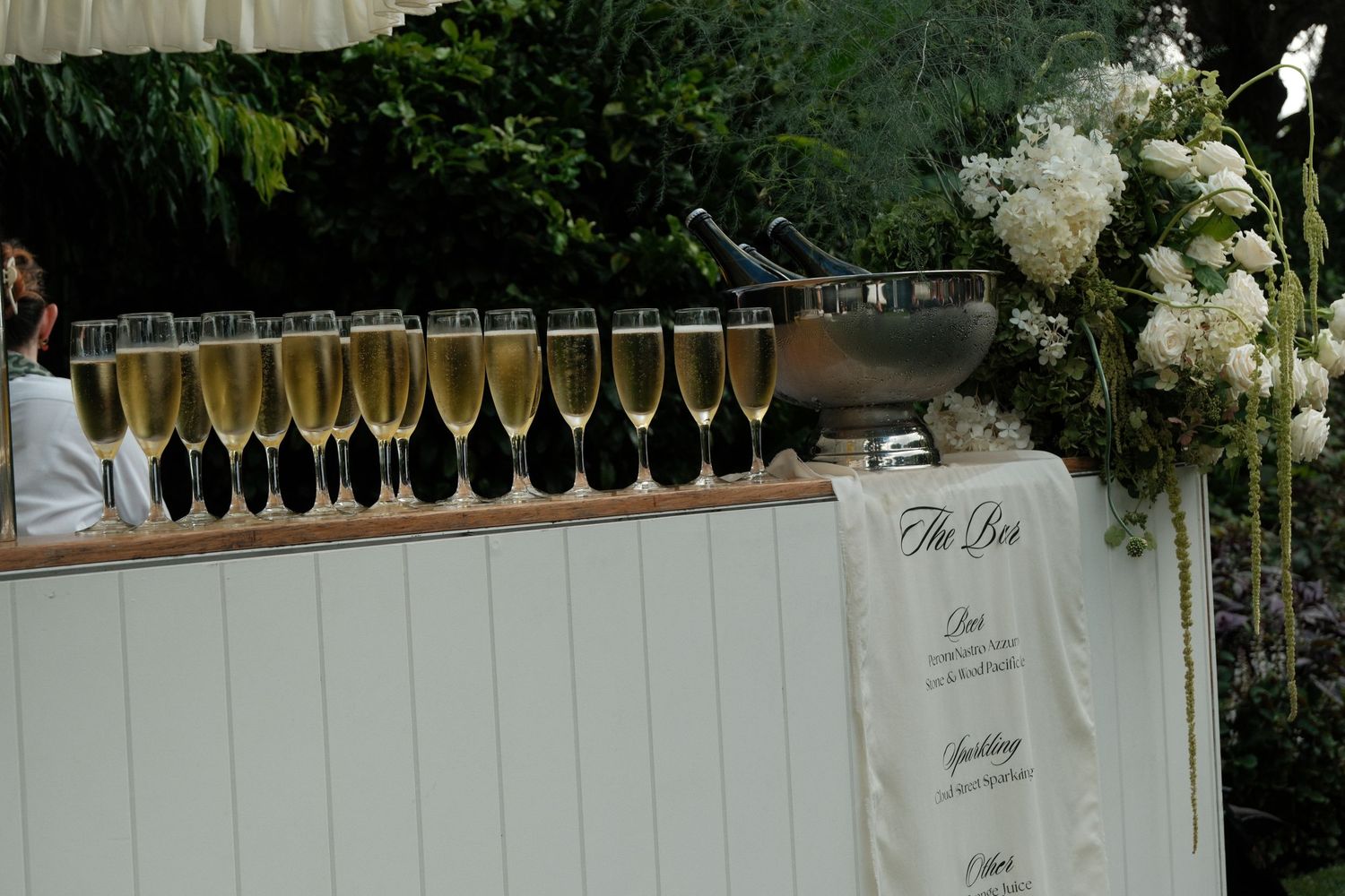 Champagne flutes lined up on a white bar with an elegant floral arrangement and menu display at an event.