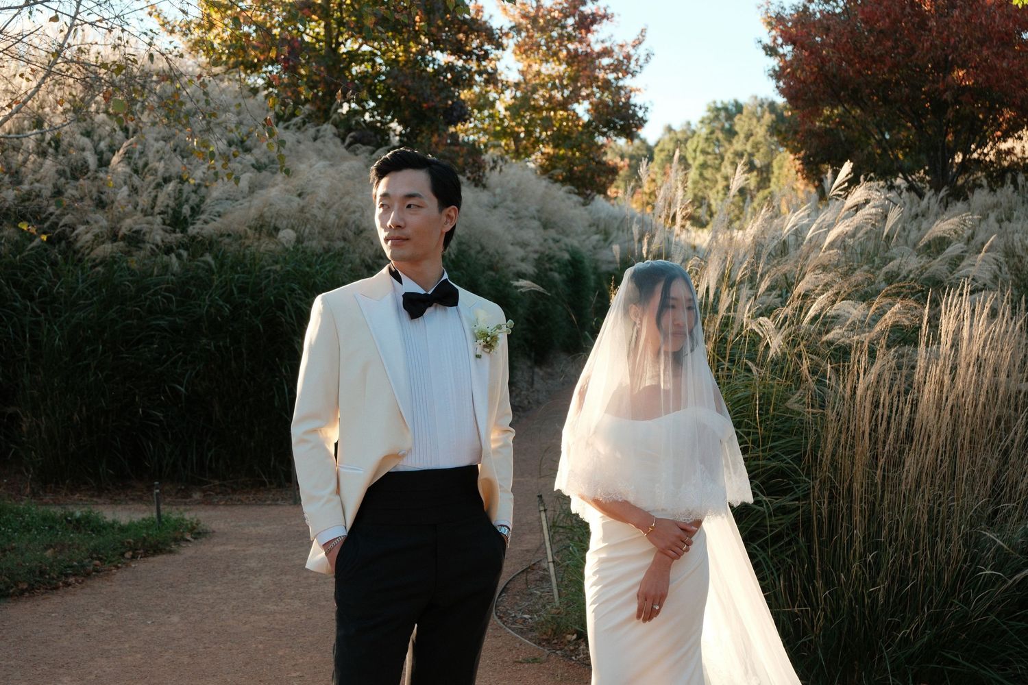 A bride in a white gown and veil stands beside a groom in a white tuxedo jacket on a garden path.