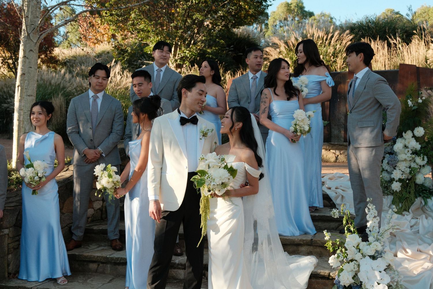 Elegant outdoor wedding ceremony with bride in white gown, groom in tuxedo, bridesmaids in blue dresses, groomsmen in gray suits.