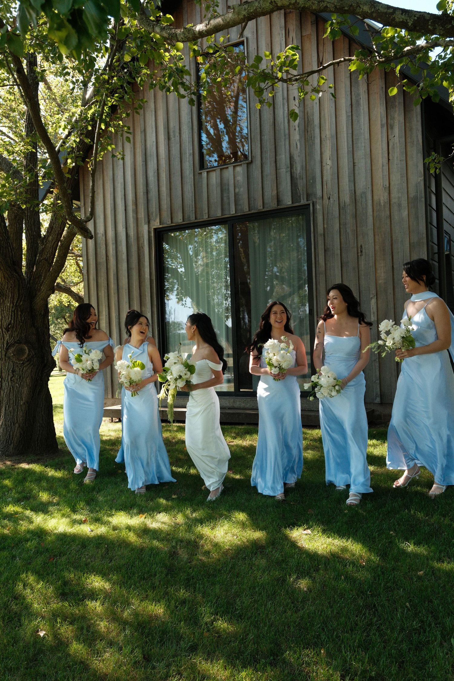 Bridesmaids in light blue gowns walk with bride in white dress carrying bouquets on green lawn near rustic barn.