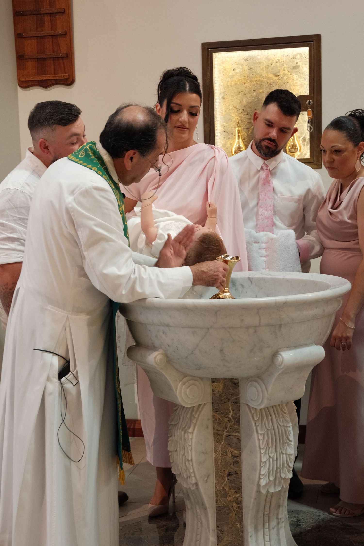 A religious baptism ceremony taking place at an ornate marble baptismal font.