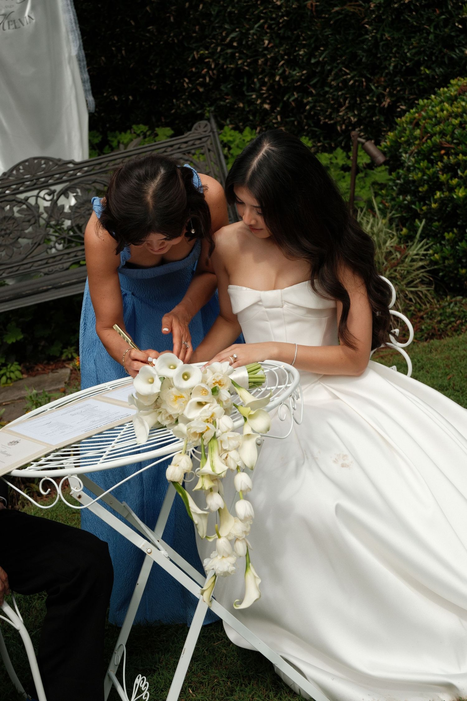 A couple in wedding attire share an intimate moment on a white garden bench with a white calla lily bouquet.